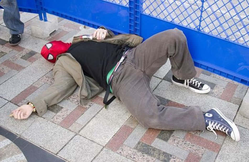 搞笑 桌布 壁紙 — 一名戴著紅色蜘蛛面具的人昏迷在磁磚人行道上，旁邊有一個藍色塑膠圍欄。