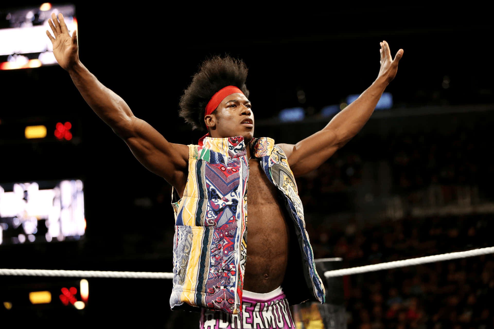 WWE 桌布 壁紙 — 一名戴著紅色頭帶、身穿彩色背心的摔角手在明亮的場館燈光下，在擂台上勝利地高舉雙臂。
