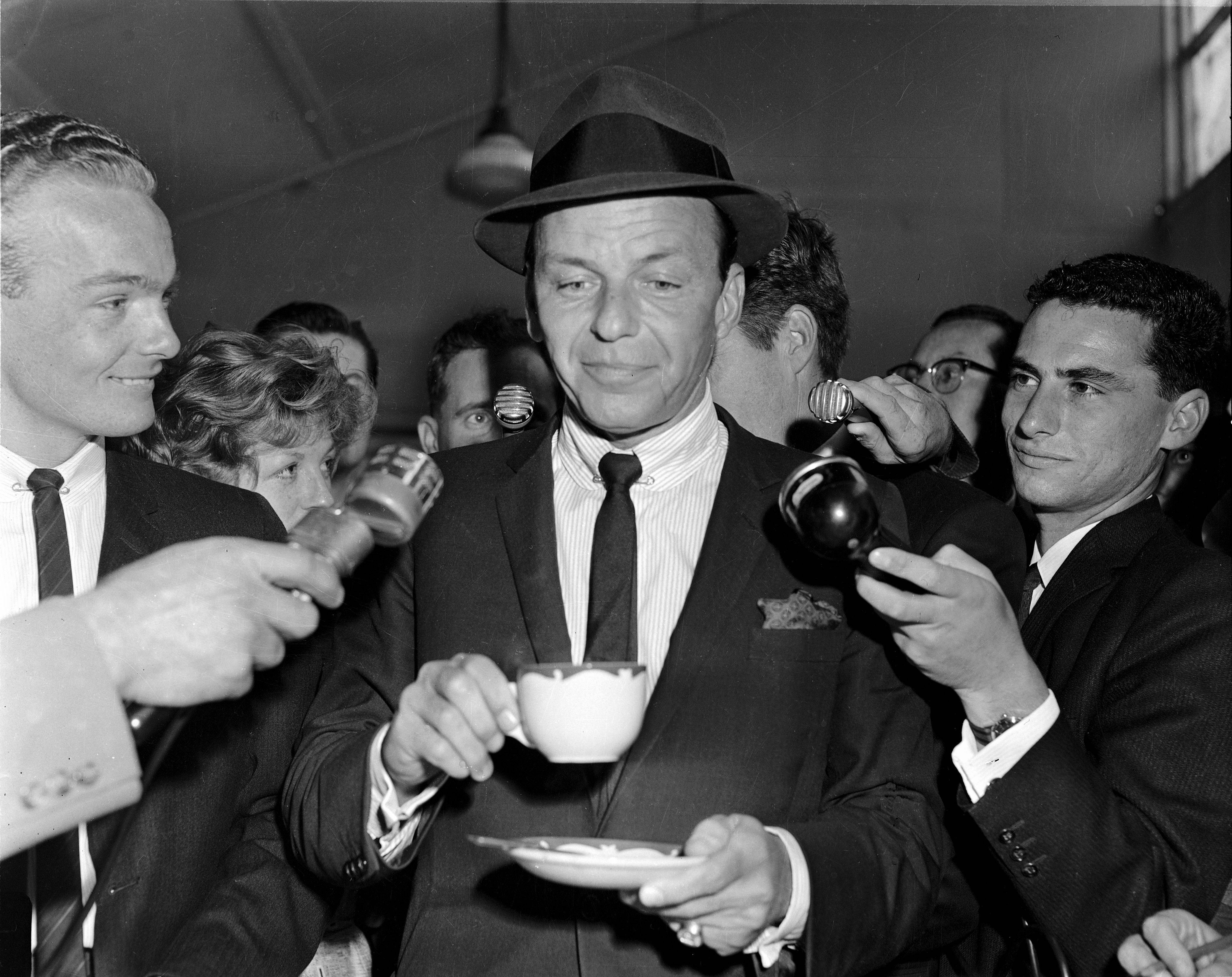 Sinatra 桌布 壁紙 — 一名戴著紳士帽、身穿西裝的男子在擠滿記者的室內環境中冷靜地喝著咖啡，記者們手持麥克風包圍著他。