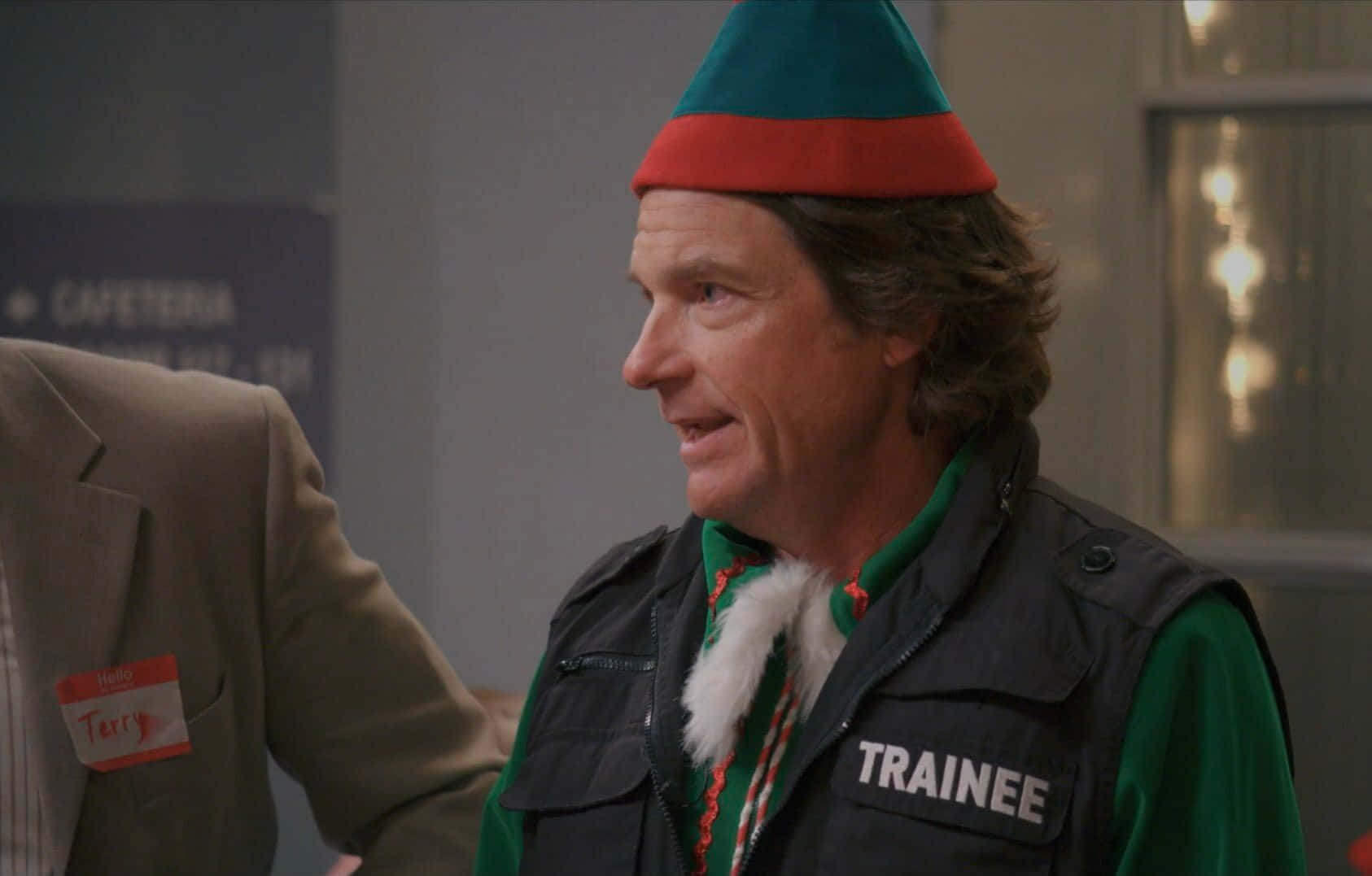 Jason Bateman 桌布 壁紙 — 一名戴著綠色和紅色精靈帽及實習生背心的男子在自助餐廳環境中側視。