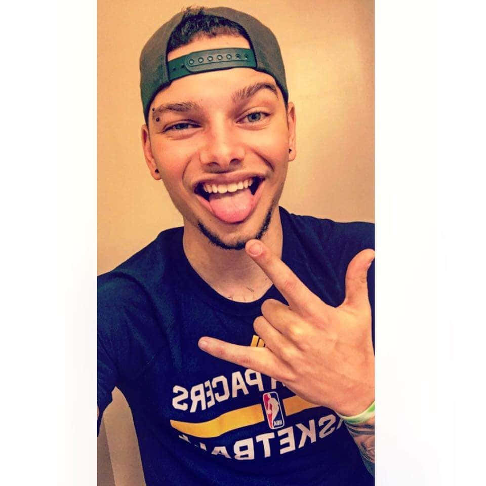 Kane Brown 桌布 壁紙 — 一名戴著綠色帽子和籃球衫的年輕男子伸出舌頭，做出手勢，微笑著。