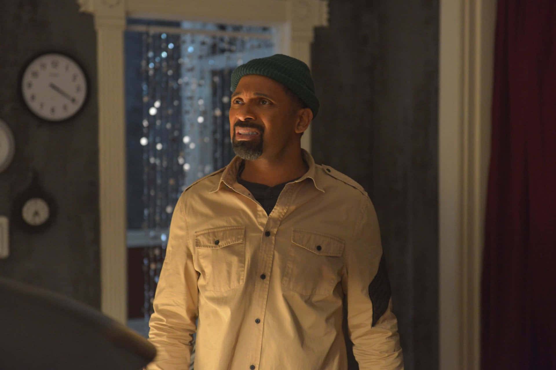 Mike Epps 桌布 壁紙 — 一名戴著綠色毛帽、身穿米色襯衫的男子站在光線昏暗的房間裡，身後有時鐘和窗簾。