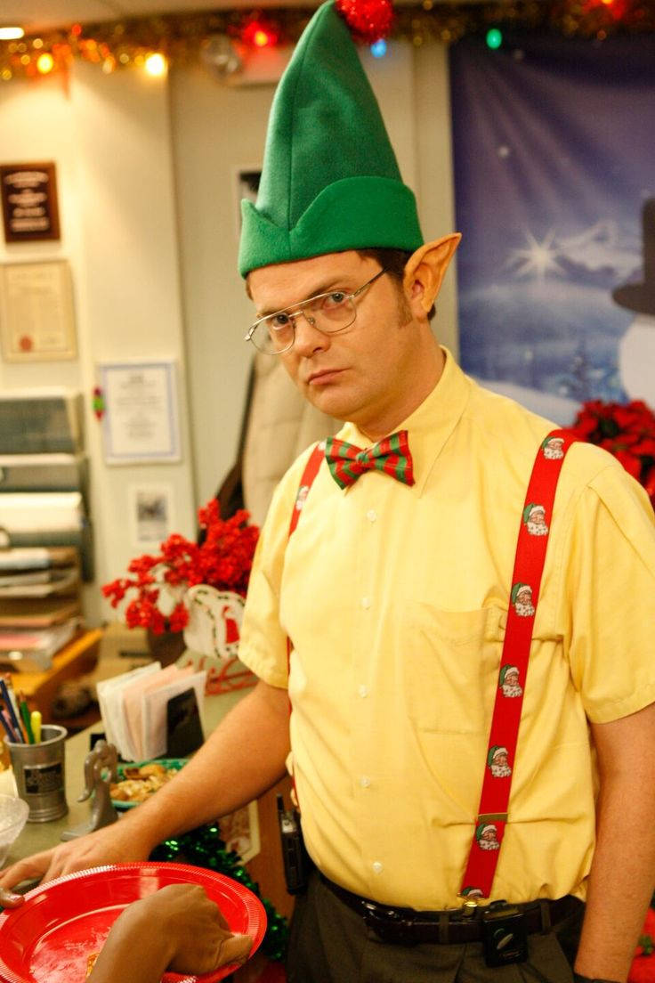 the office christmas 桌布 壁紙 — 一名戴著綠色精靈帽和紅色吊帶的男人站在一個裝飾著聖誕燈飾和聖誕紅的節日辦公室裡。