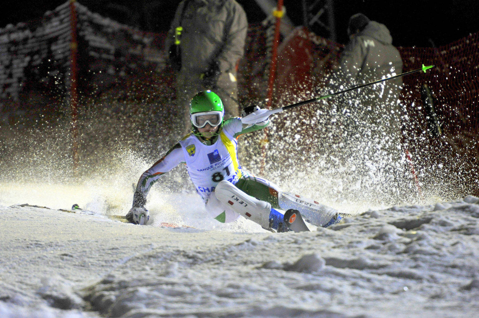 Olympics 桌布 壁紙 — 一名戴著綠色頭盔的滑雪者在夜間穿過雪地，在明亮的燈光下將粉雪噴灑到空中。