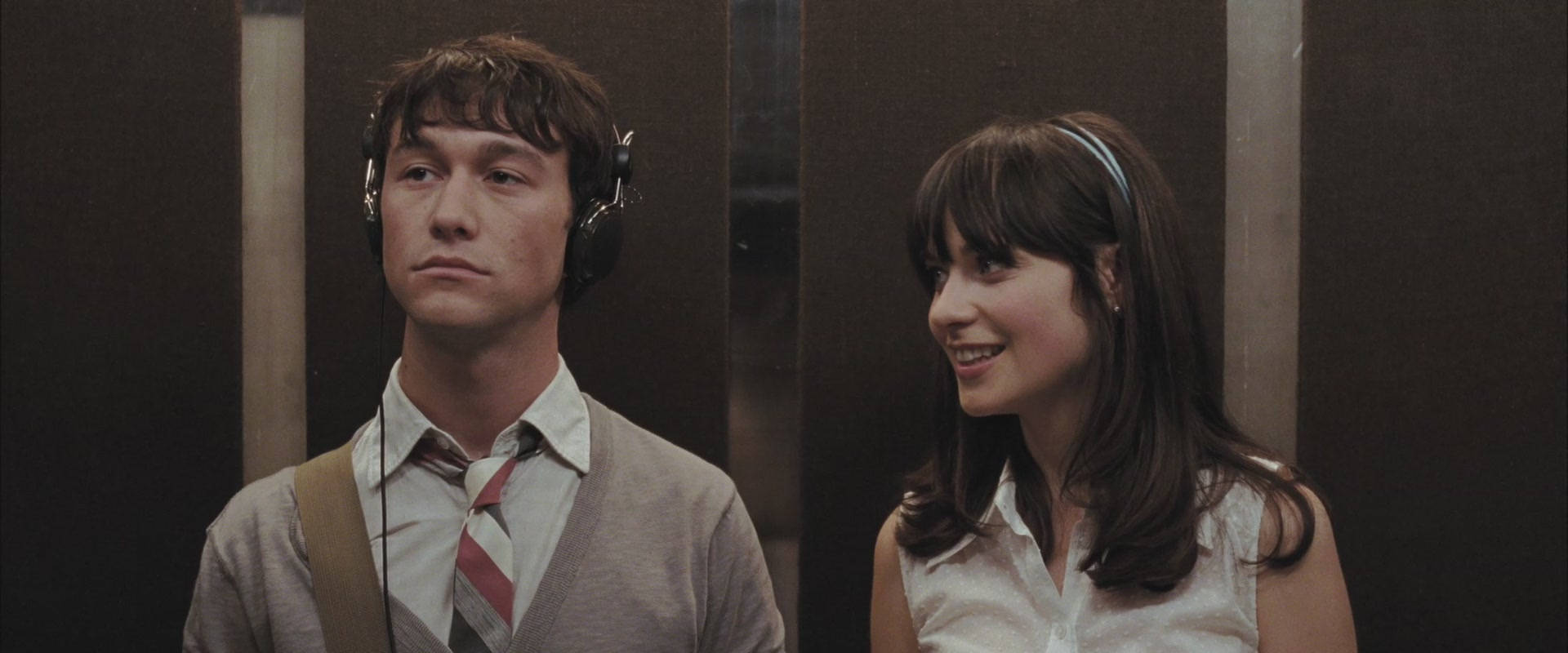 500 Days Of Summer 桌布 壁紙 — 一名戴著耳機的男子和一名微笑的女子並肩站在光線昏暗、金屬牆壁的電梯內。