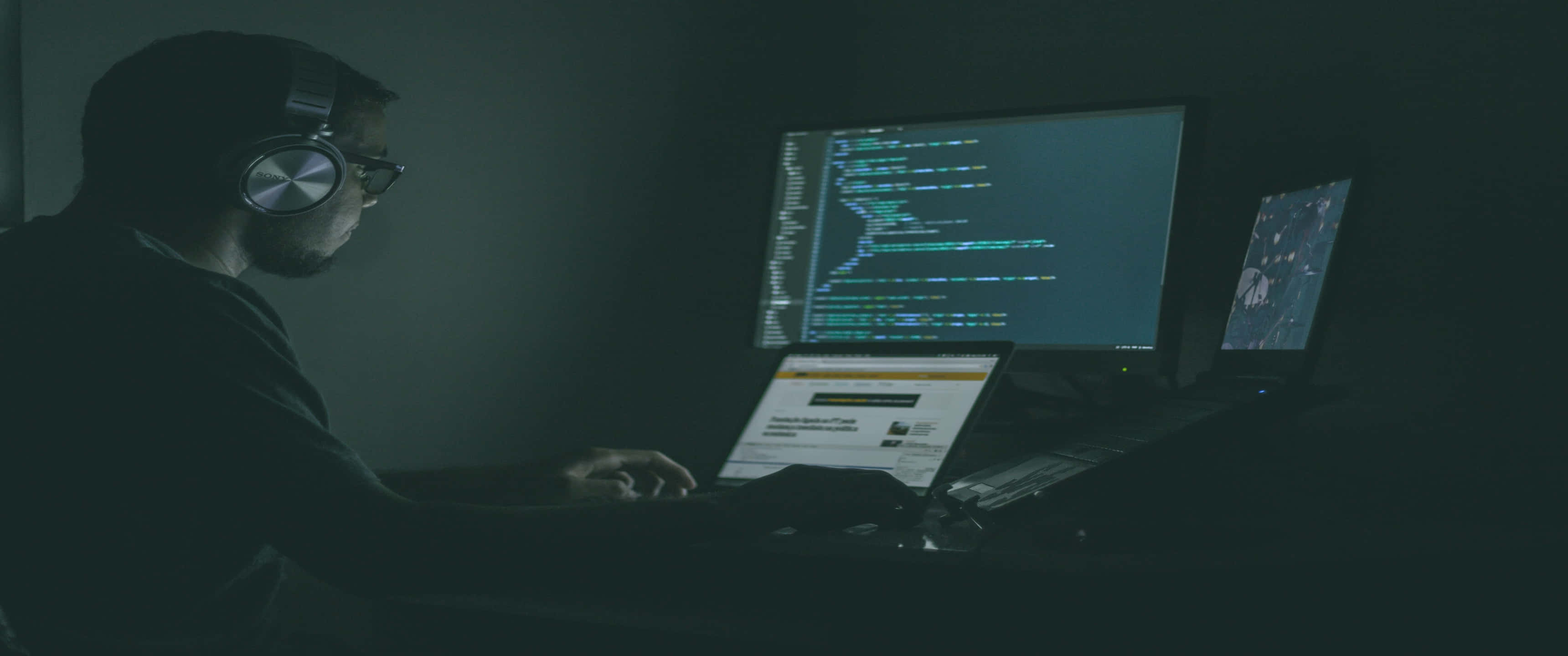 3440x1440p Programming 桌布 壁紙 — 一名戴著耳機的程式設計師在昏暗的書桌前專注工作，周圍是顯示程式碼的發光電腦螢幕。