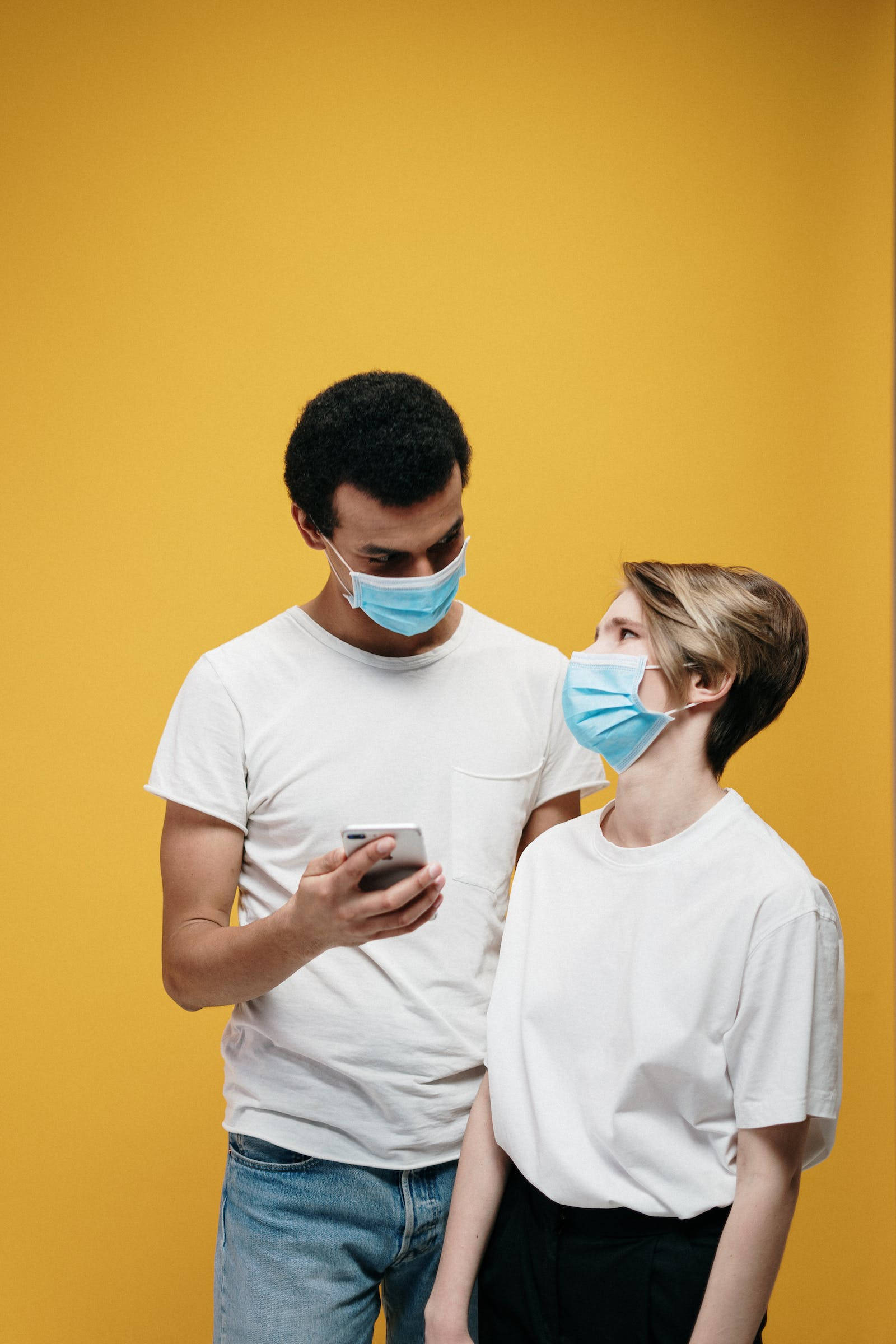 Love iPhone 桌布 壁紙 — 一名戴著藍色外科口罩的男女傾身觀看智慧型手機，背景為純芥末黃色。