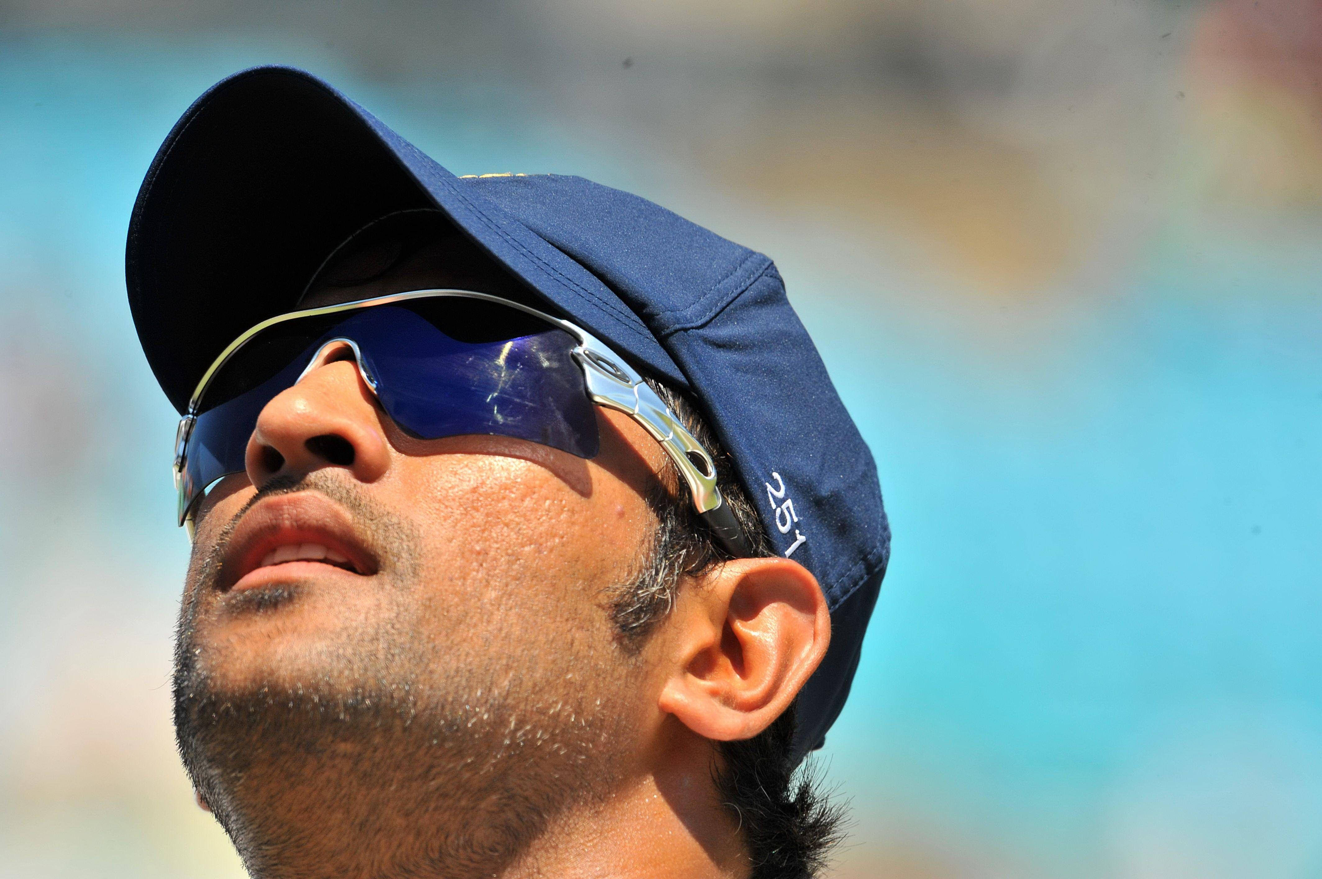 Ms Dhoni 桌布 壁紙 — 一名戴著藍色帽子和太陽眼鏡的男子，在明亮的藍天下向上凝視的特寫。