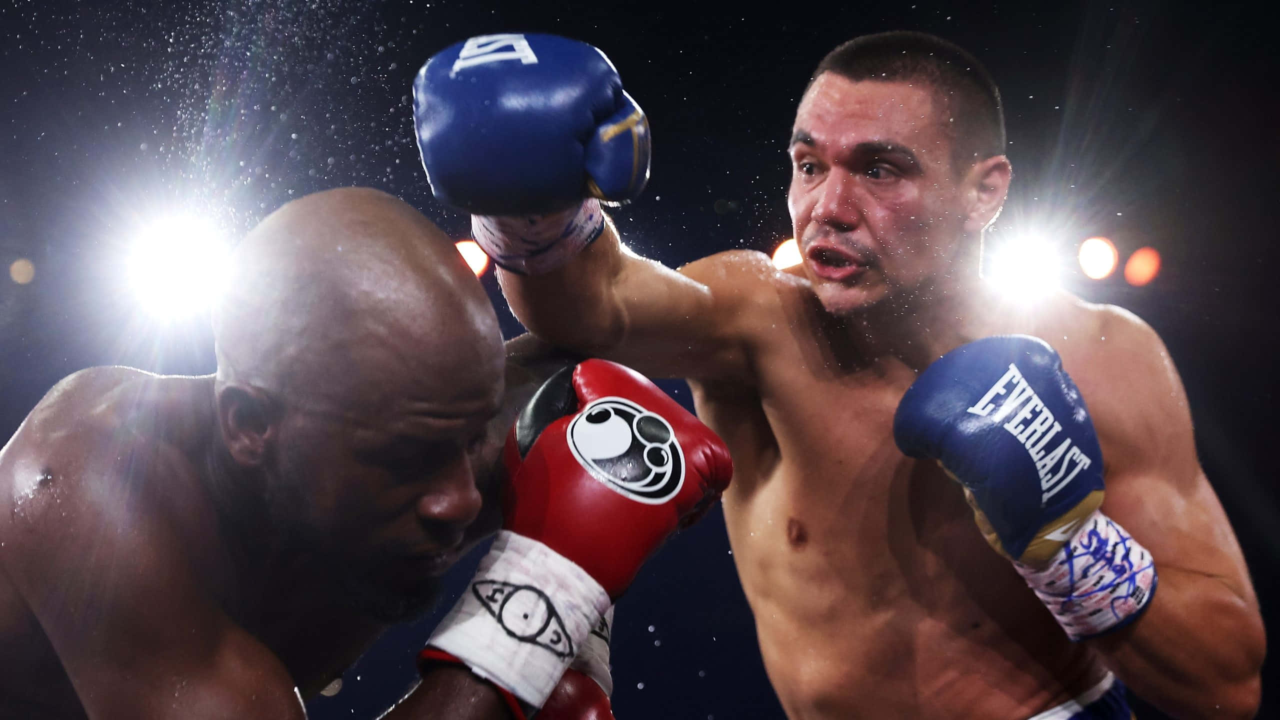 Tim Tszyu 桌布 壁紙 — 一名戴著藍色手套的拳擊手在燈光明亮的擂台上向對手打出強力一拳，汗水飛濺。