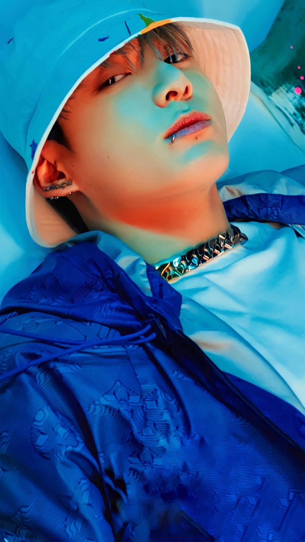 BTS 桌布 壁紙 — 一名戴著藍色漁夫帽和連帽衫的年輕男子，在霓虹藍光下戴著金屬鏈條項鍊向後躺。