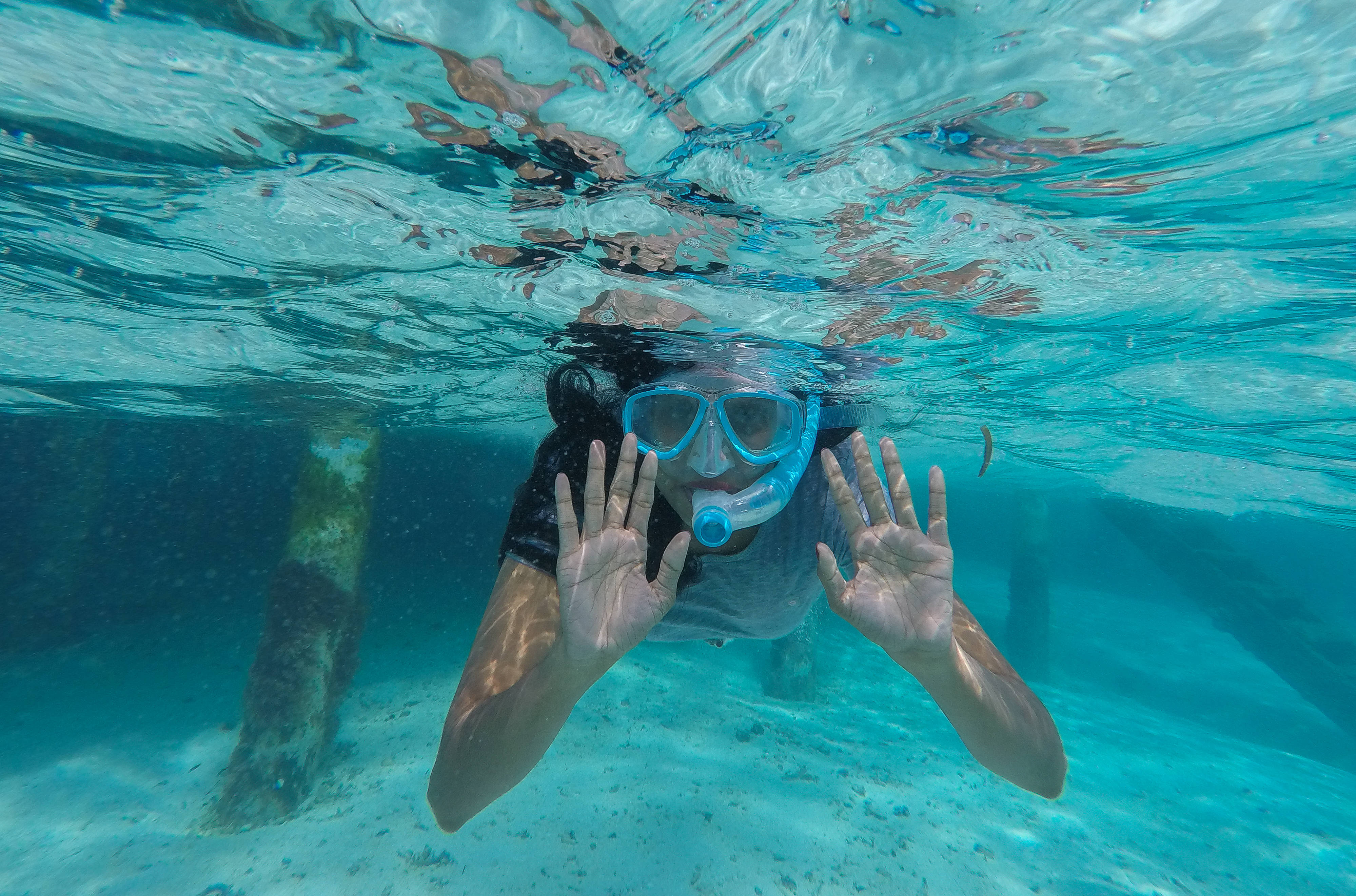 Snorkeling 桌布 壁紙 — 一名戴著藍色裝備的浮潛者在陽光照射的廢墟附近、清澈碧綠的水中揮手。