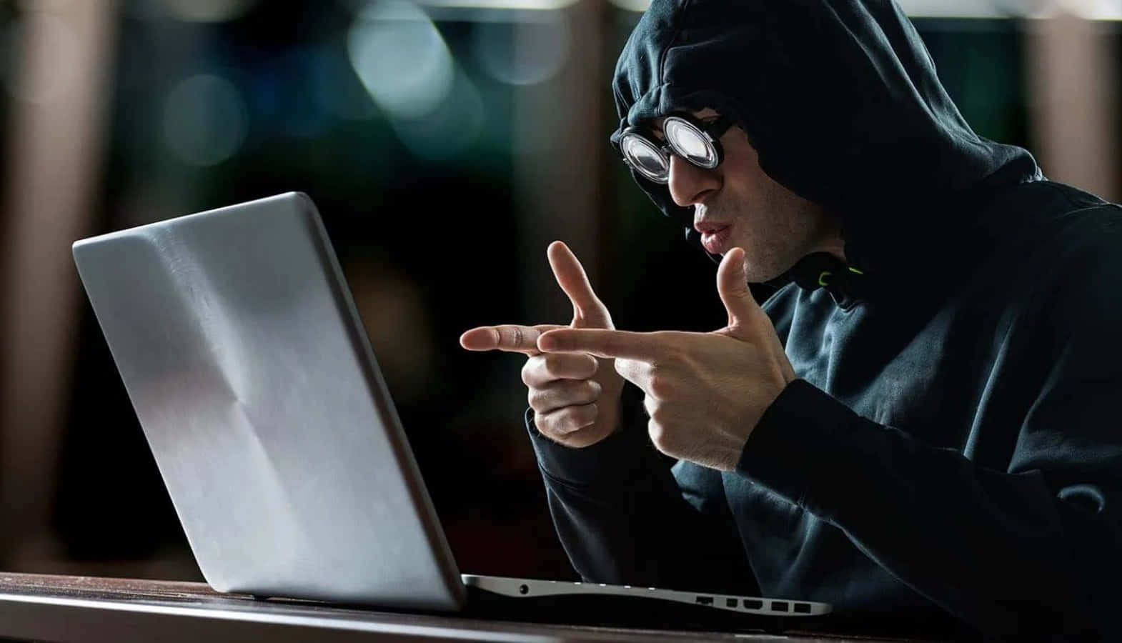 黑客 桌布 壁紙 — 一名戴著護目鏡的連帽駭客在昏暗的房間中指向筆記型電腦螢幕，背景燈光模糊。