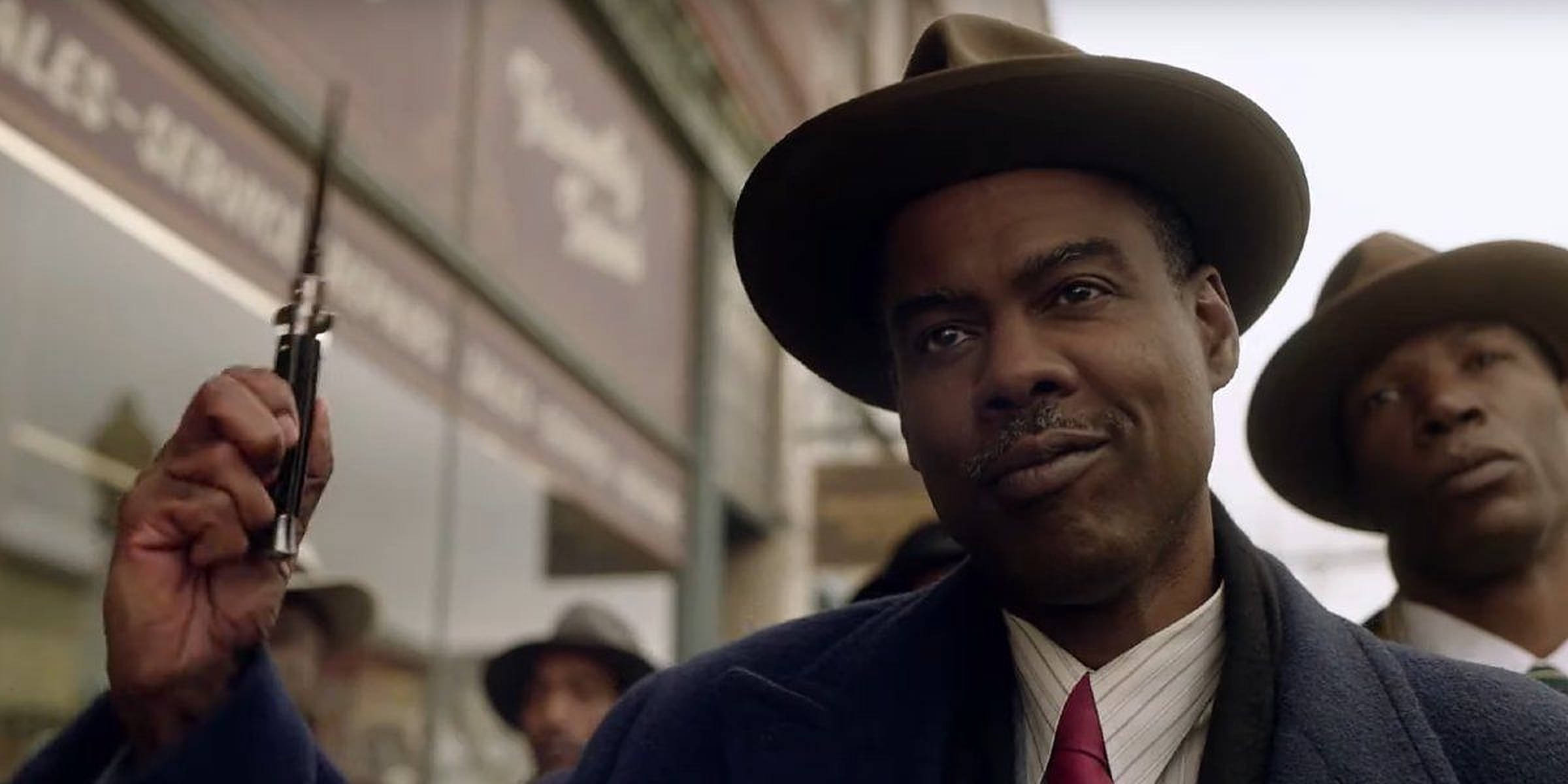 Chris Rock 桌布 壁紙 — 一名戴著費多拉帽、身穿西裝的男子站在城市街道上，手持手槍，背景中有另一名男子。