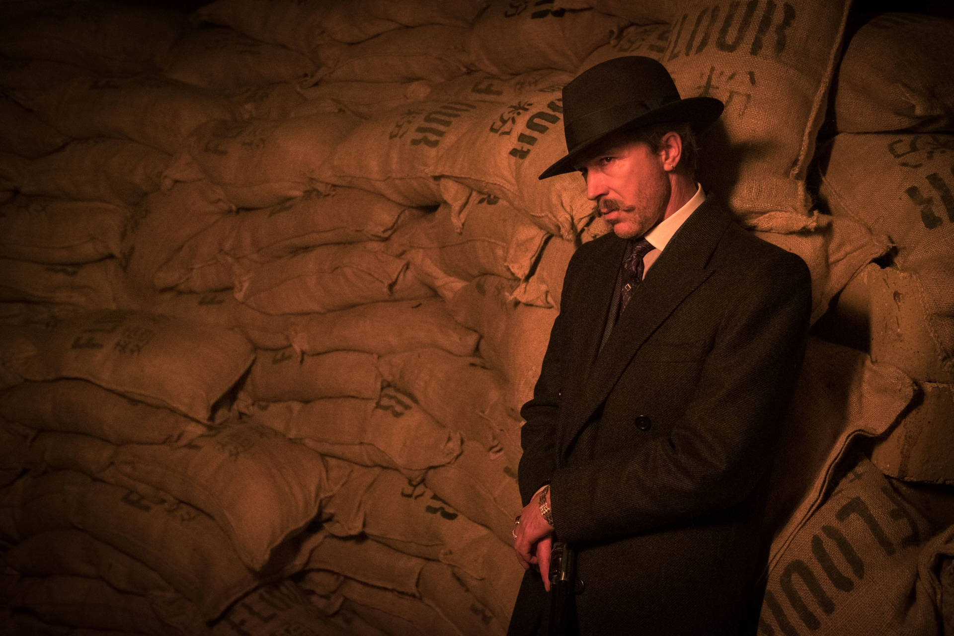 戴軟呢帽穿西裝的男子，在昏暗空間中靠著粗麻布袋牆。 — Peaky Blinders 壁紙