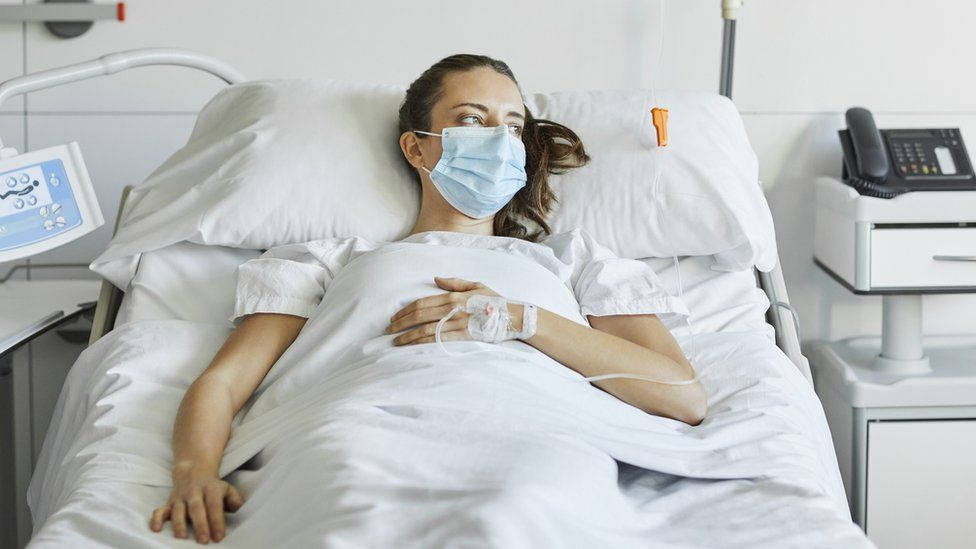 Mask 桌布 壁紙 — 一名戴著醫用口罩的女性躺在醫院的床上，正在接受靜脈輸液，周圍是醫療設備和白色床單。