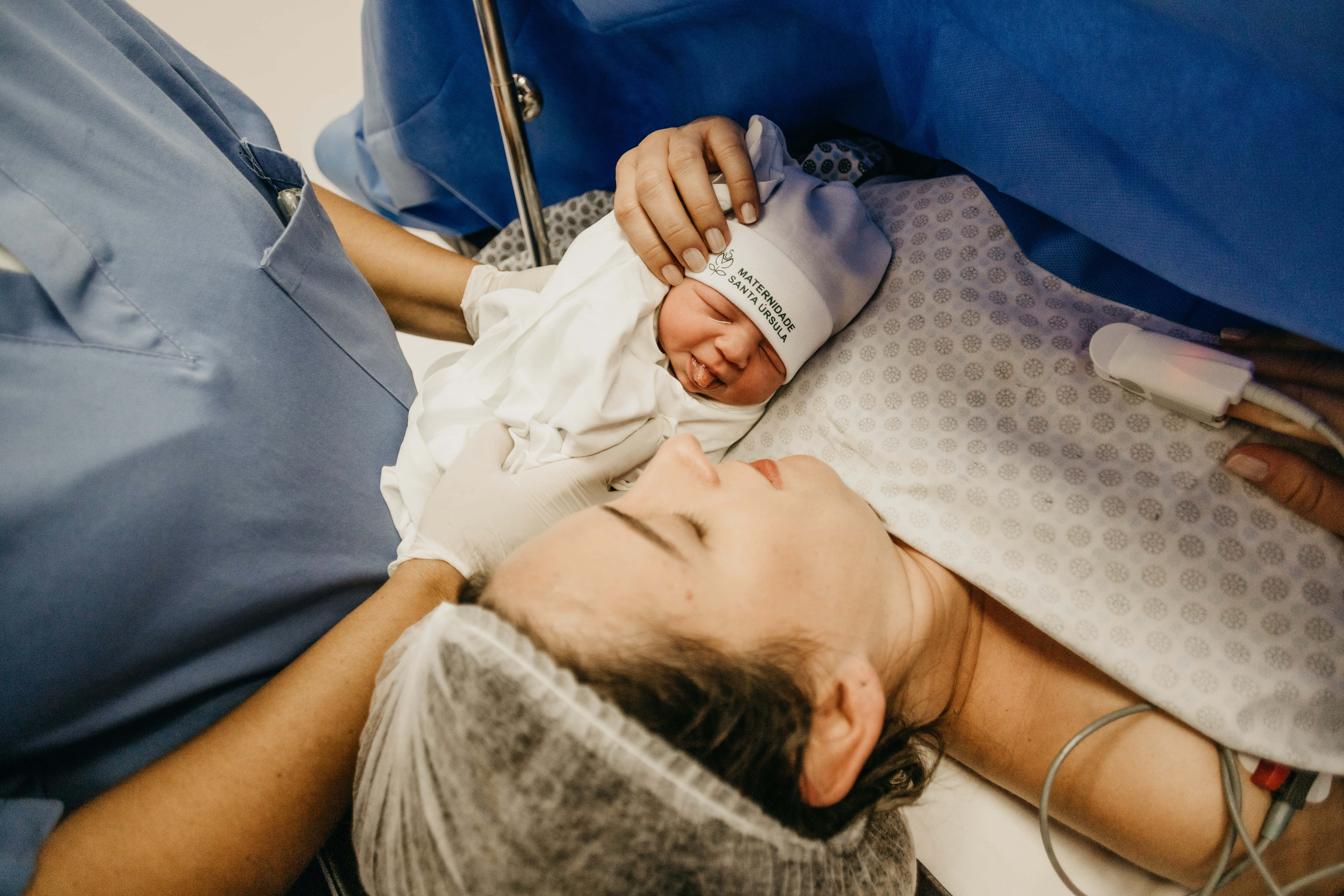 Mom 桌布 壁紙 — 一名戴著醫院帽的新生嬰兒在產房內，躺在母親的胸口上休息，周圍有醫護人員和設備。
