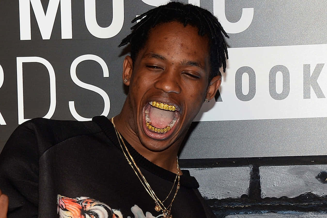 Travis Scott Funny 桌布 壁紙 — 一名戴著金色牙套和辮子頭的男性在音樂頒獎活動上大笑，身穿圖案T恤和疊戴金鍊。