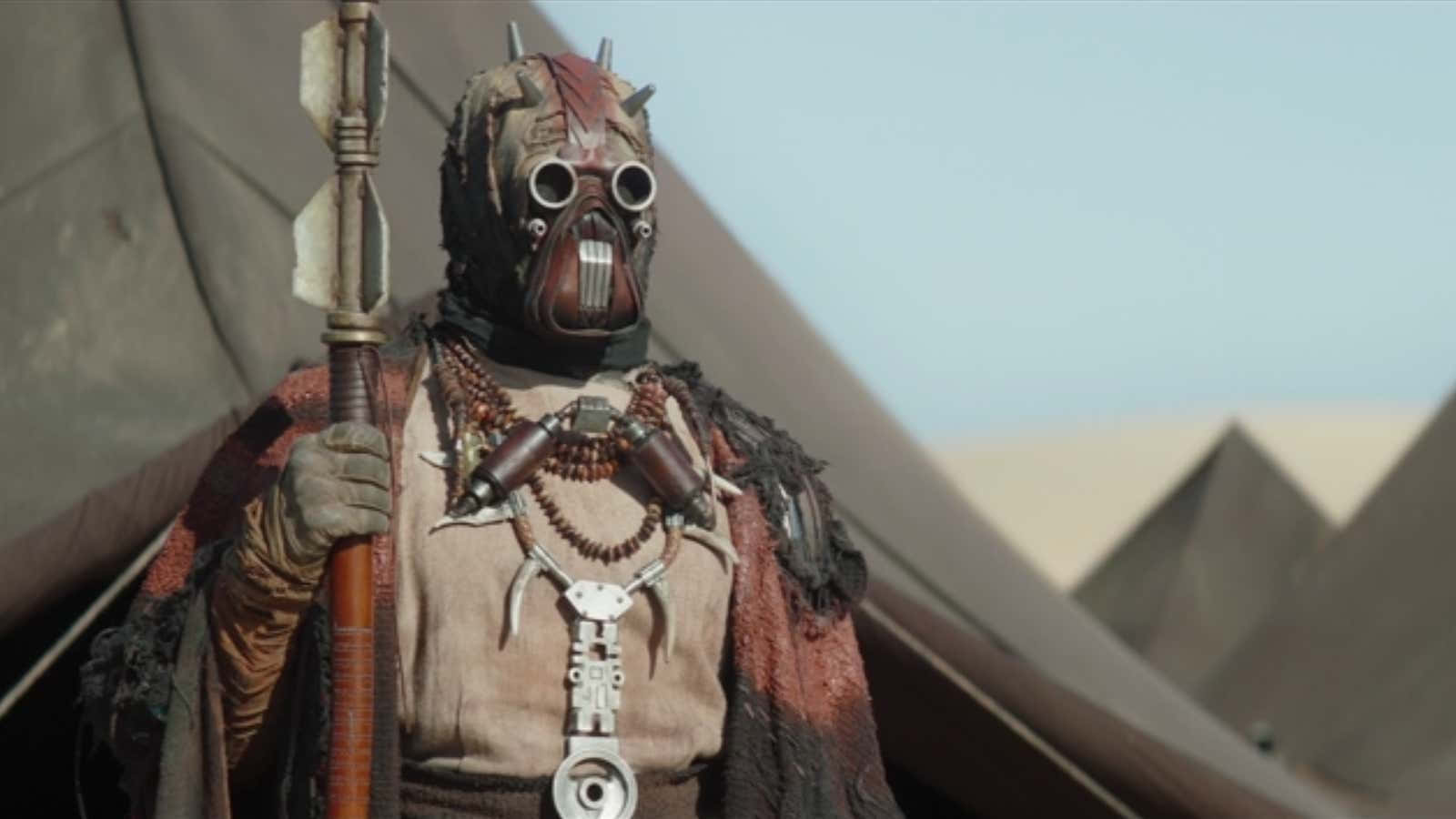 Tusken Raiders 桌布 壁紙 — 一名戴著面具的戰士身穿沙漠裝備，在晴朗的天空下手持長矛，背景有帳篷狀結構。