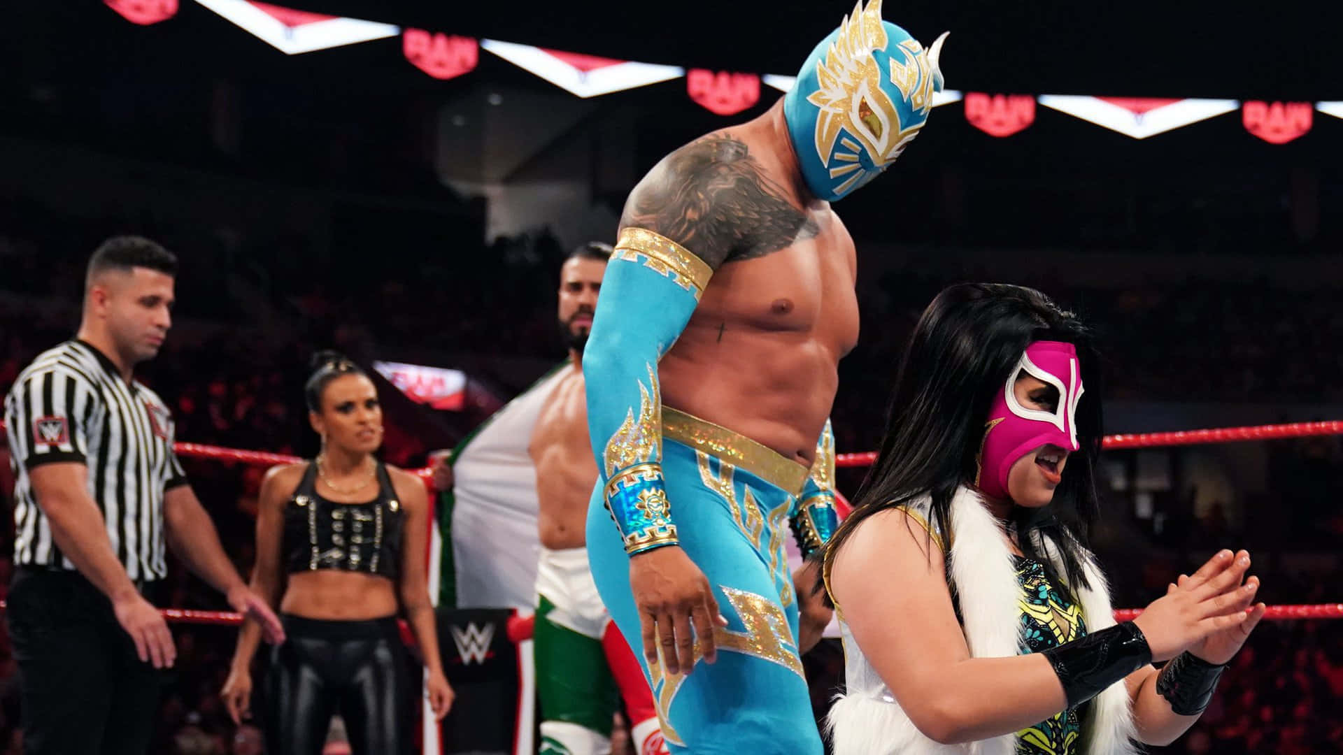 WWE 桌布 壁紙 — 一名戴著面具的摔角手身穿藍色和金色裝備，在擂台上高高站立，而戴著面具的夥伴在場館燈光下在他旁邊鼓掌。