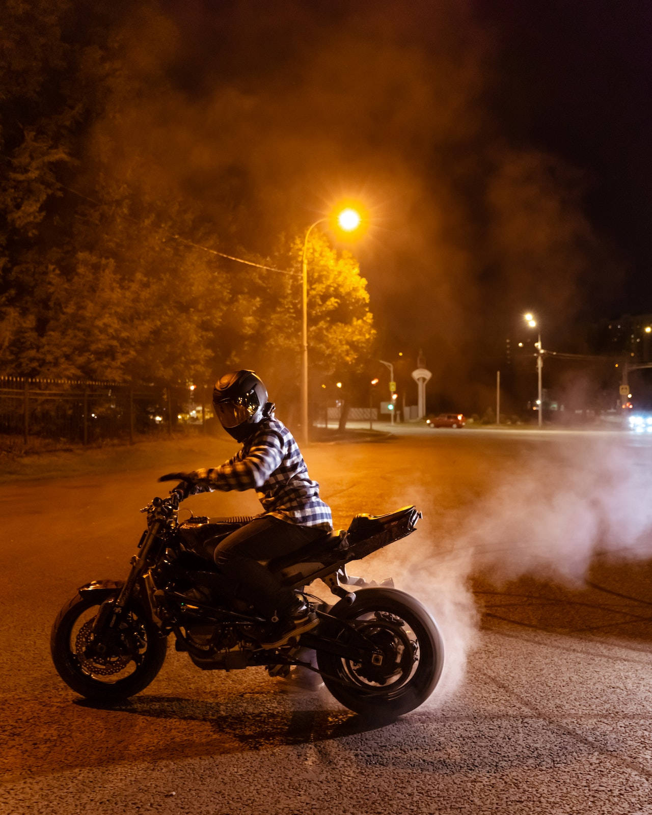 Bike Rider 桌布 壁紙 — 一名戴著頭盔、身穿格紋襯衫的摩托車手在夜晚燒胎，在發光的街燈下揚起煙霧。