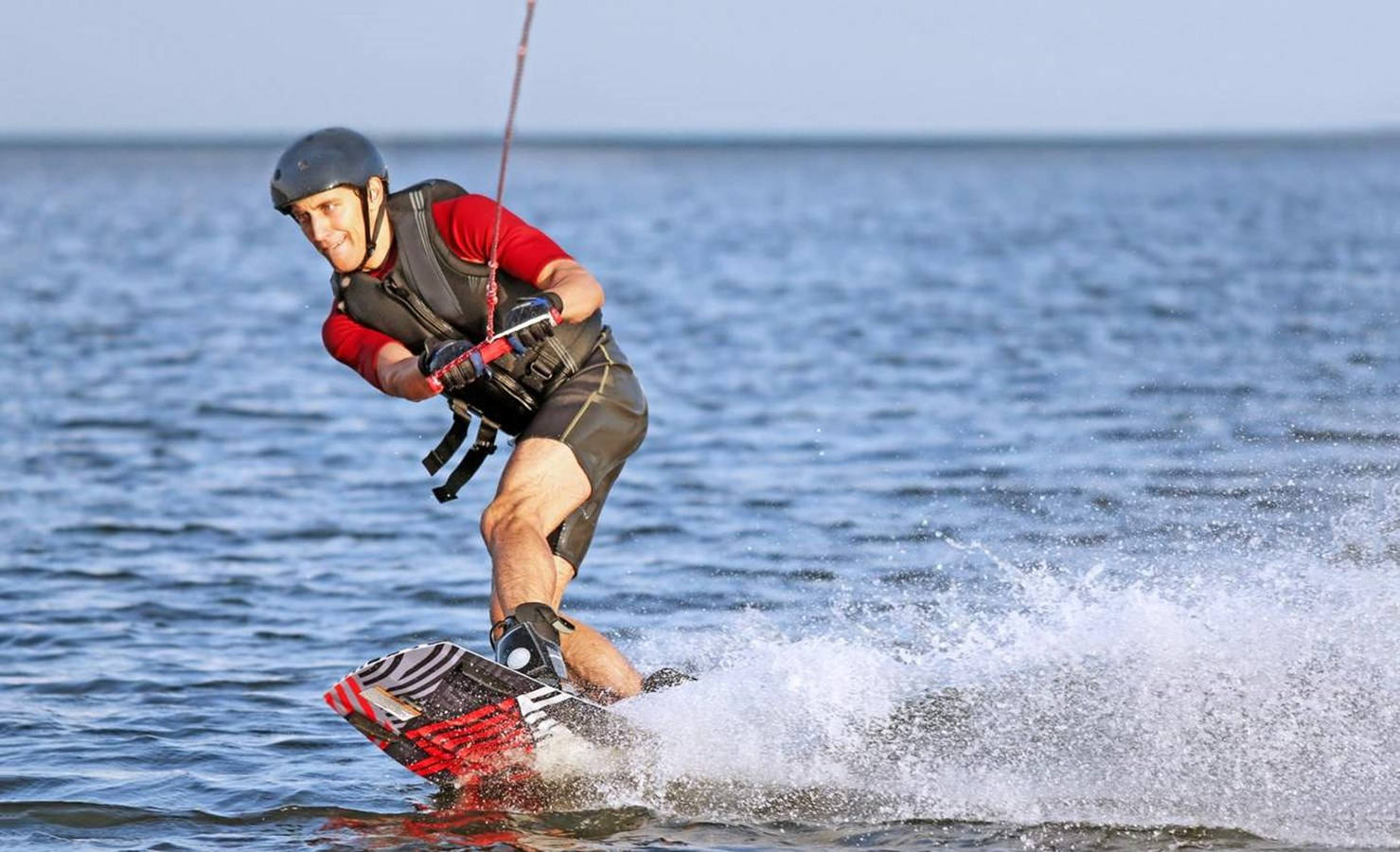 surfing 桌布 壁紙 — 一名戴著頭盔和救生衣的男子在平靜的藍色湖上玩滑水板，微笑著，身後濺起水花。
