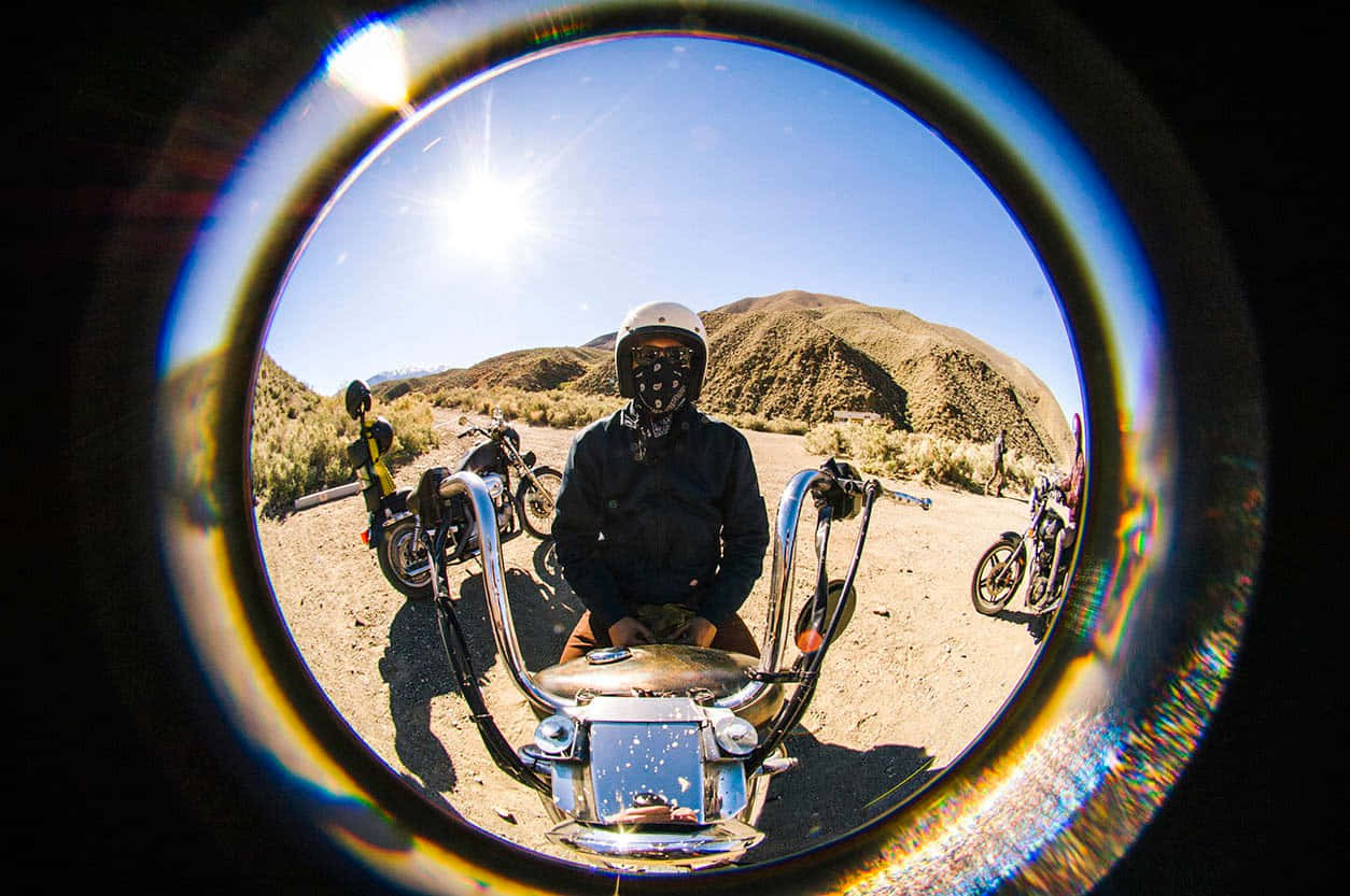 Fisheye 桌布 壁紙 — 一名戴著頭盔和面罩的摩托車手坐在一輛鍍鉻摩托車上，透過魚眼鏡頭觀看，背景有明亮的太陽和沙漠山丘。