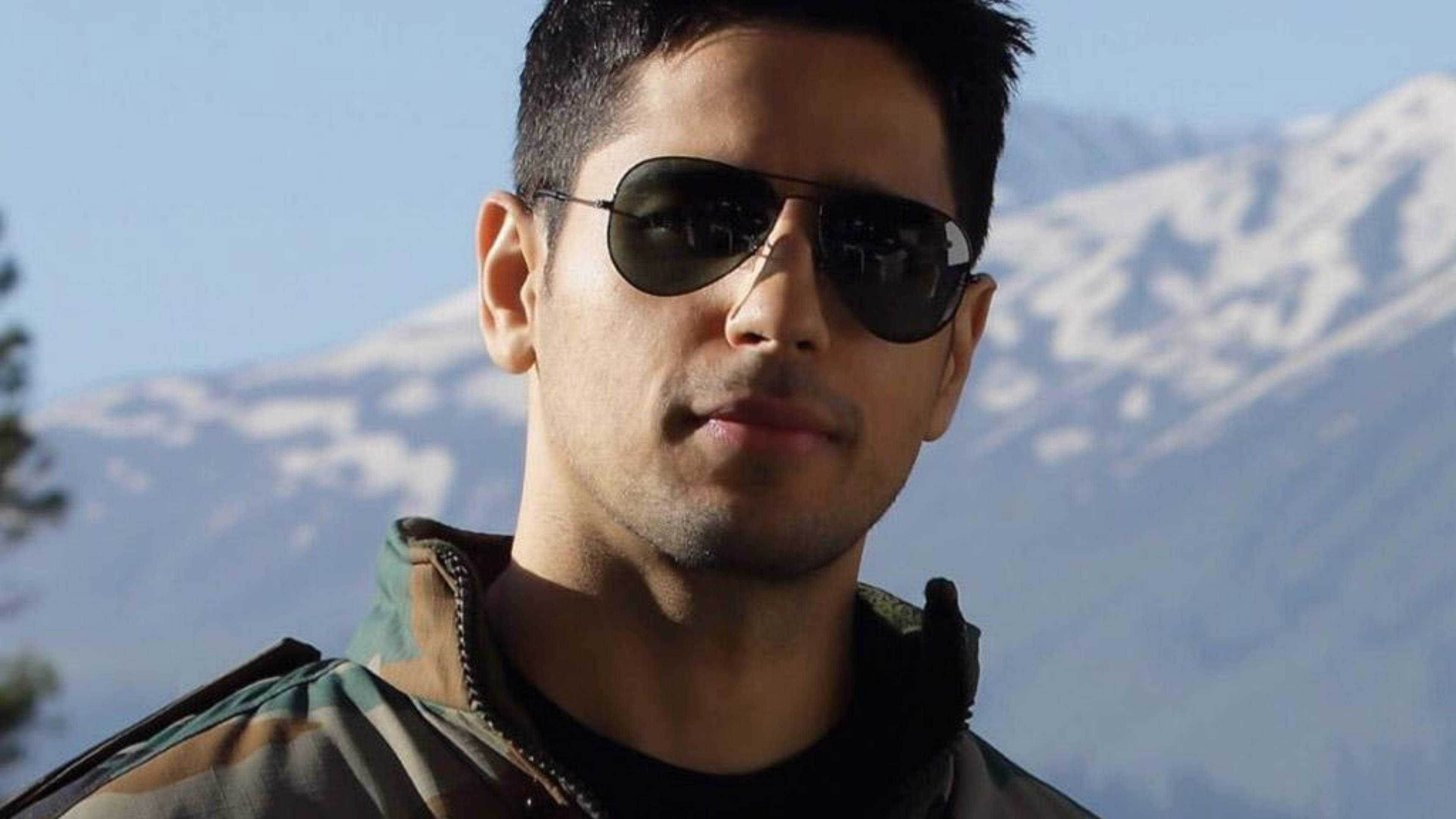 Sidharth Malhotra 桌布 壁紙 — 一名戴著飛行員太陽眼鏡和迷彩外套的男子，在晴朗藍天下以積雪山脈為背景自信地站立。