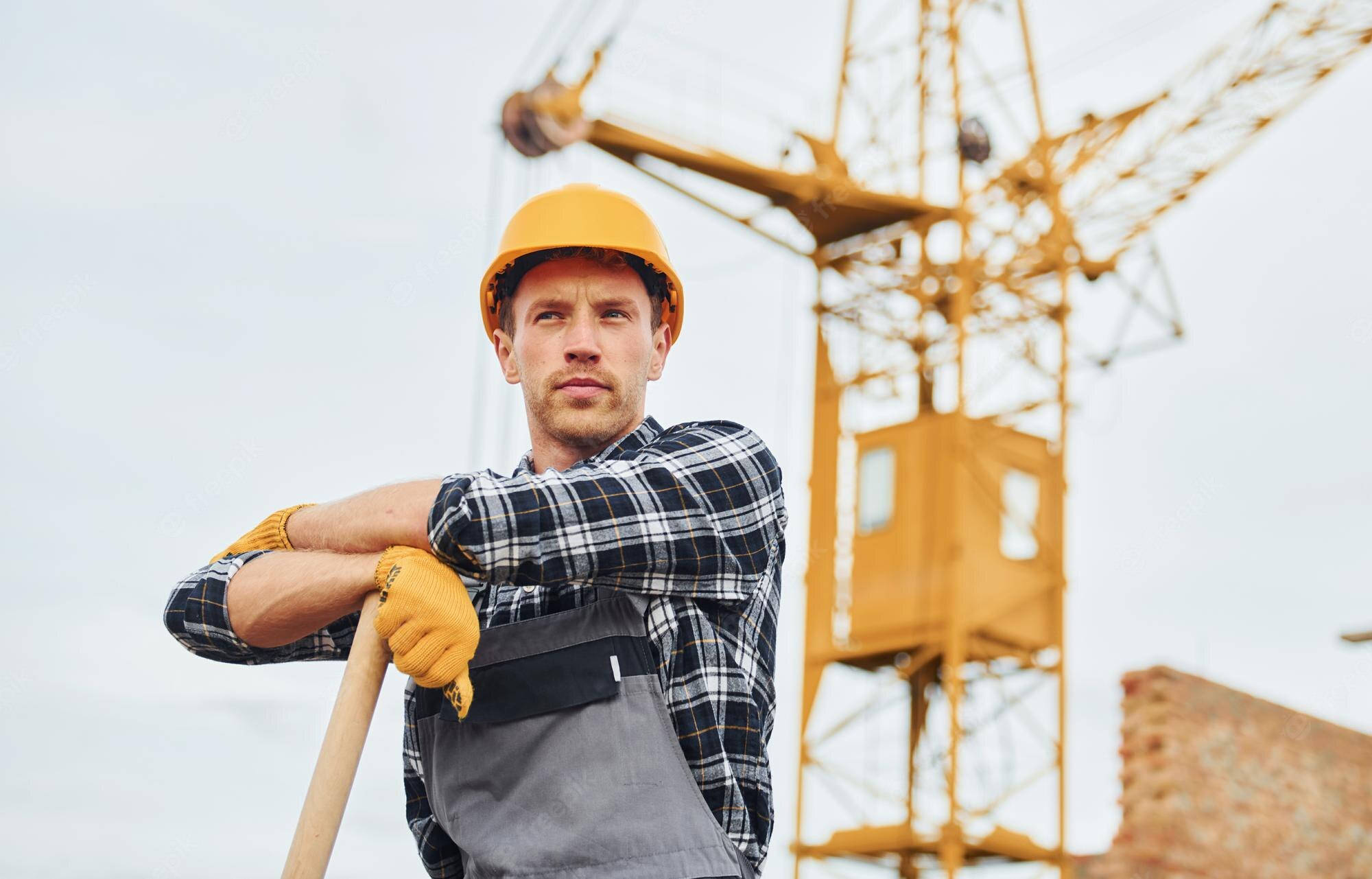 Working 桌布 壁紙 — 一名戴著黃色安全帽、穿著格紋襯衫的建築工人自信地站著，手持鏟子，背景是一台大型起重機。