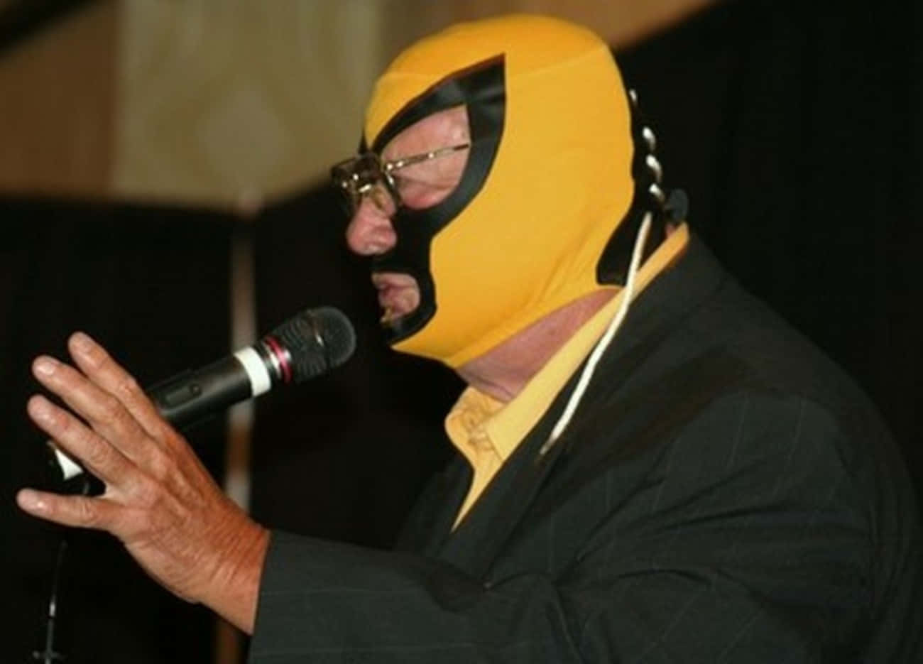 WWE 桌布 壁紙 — 一名戴著黃色自由式摔角面具和眼鏡的男子，身穿黑色西裝，對著麥克風說話。