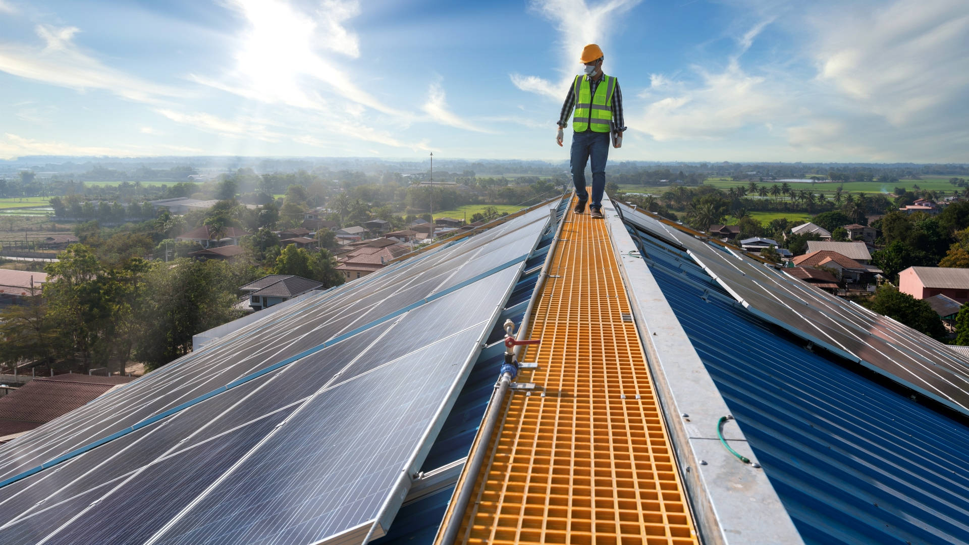 Solar Panel 桌布 壁紙 — 一名戴著黃色頭盔、穿著綠色背心的工人，在陽光明媚的屋頂上，沿著明亮的橙色走道，在成排的太陽能板之間行走，俯瞰郊區景觀。