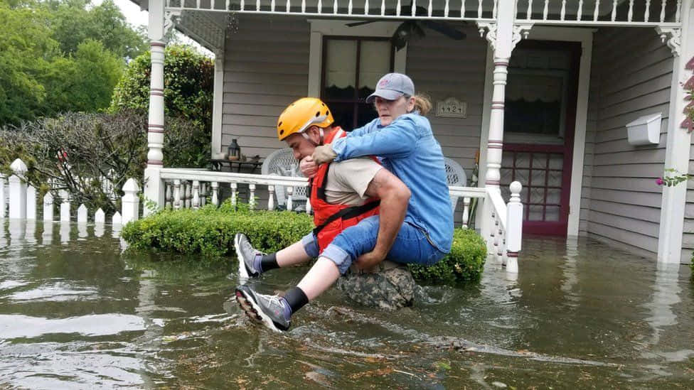 Flood 桌布 壁紙 — 一名戴著黃色頭盔的救援人員抱著一位老婦人穿過洪水，帶到被淹沒的門廊附近的安全地帶。