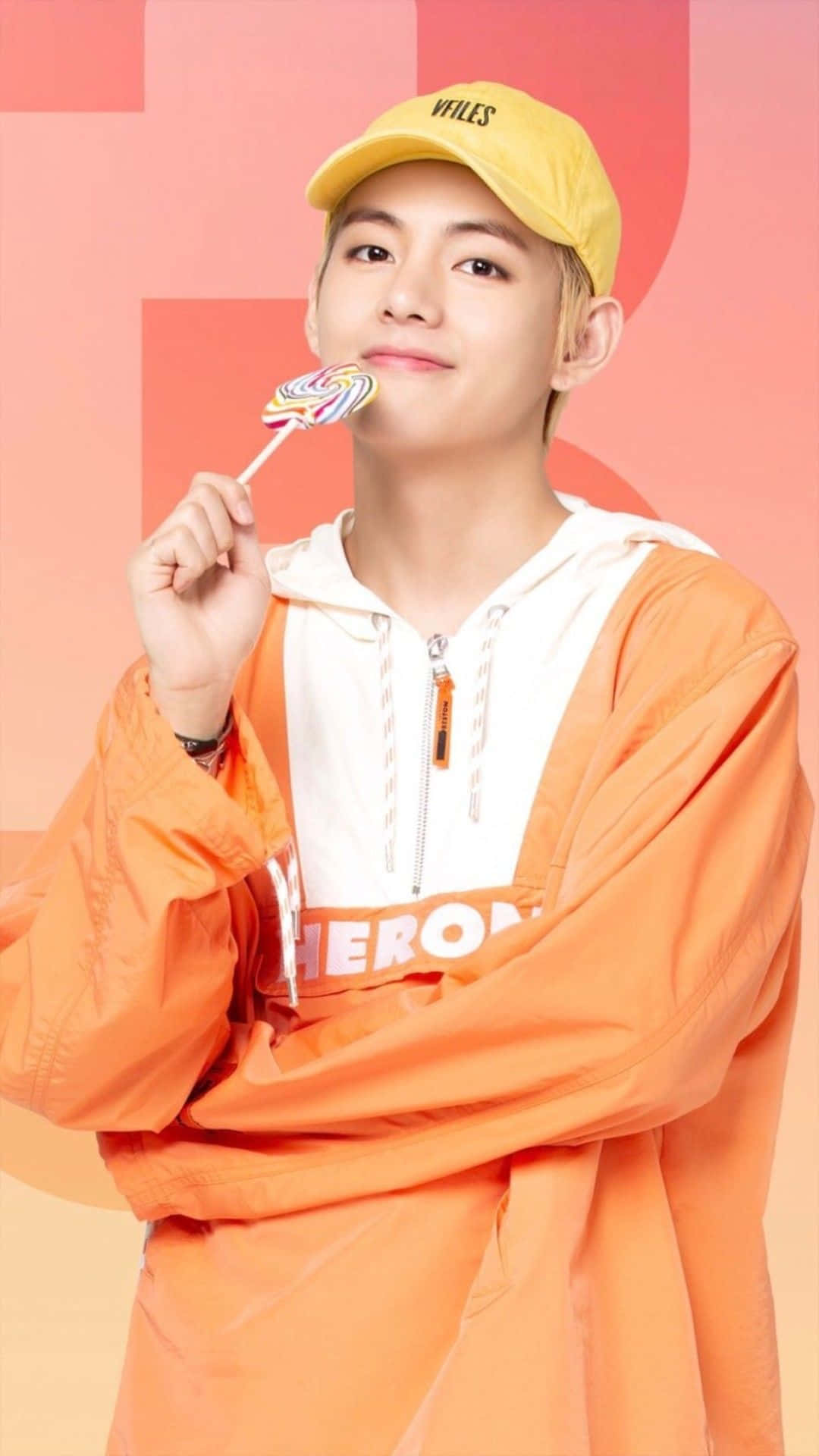 Taehyung Cute 桌布 壁紙 — 一名戴著黃色鴨舌帽、身穿橙色連帽上衣的年輕男子，在柔和的粉紅色幾何背景前拿著一支彩色棒棒糖。