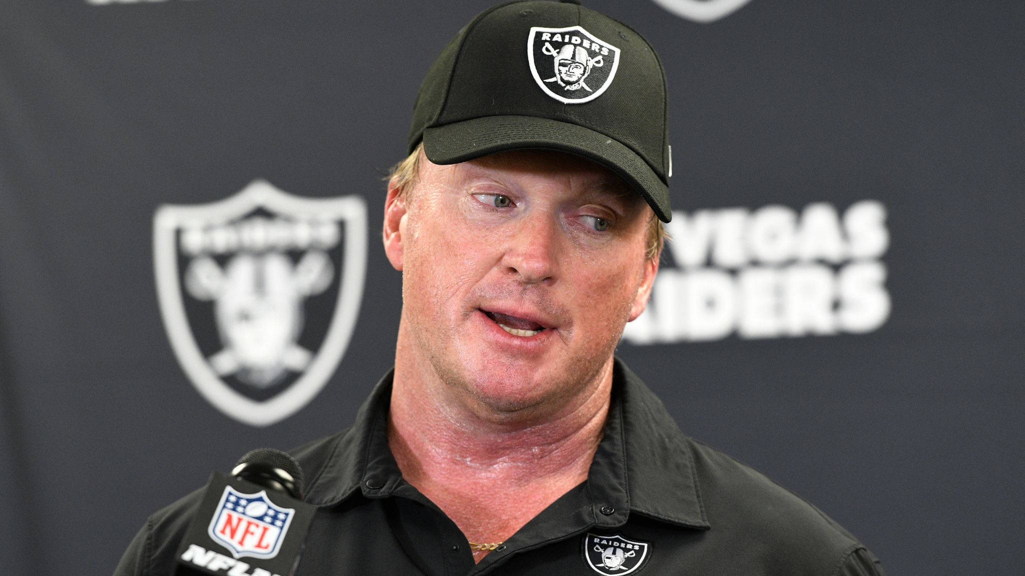 運動 桌布 壁紙 — 一名戴著黑色Raiders帽子和穿著黑色Raiders襯衫的男子在記者會上發言，身後有球隊標誌。