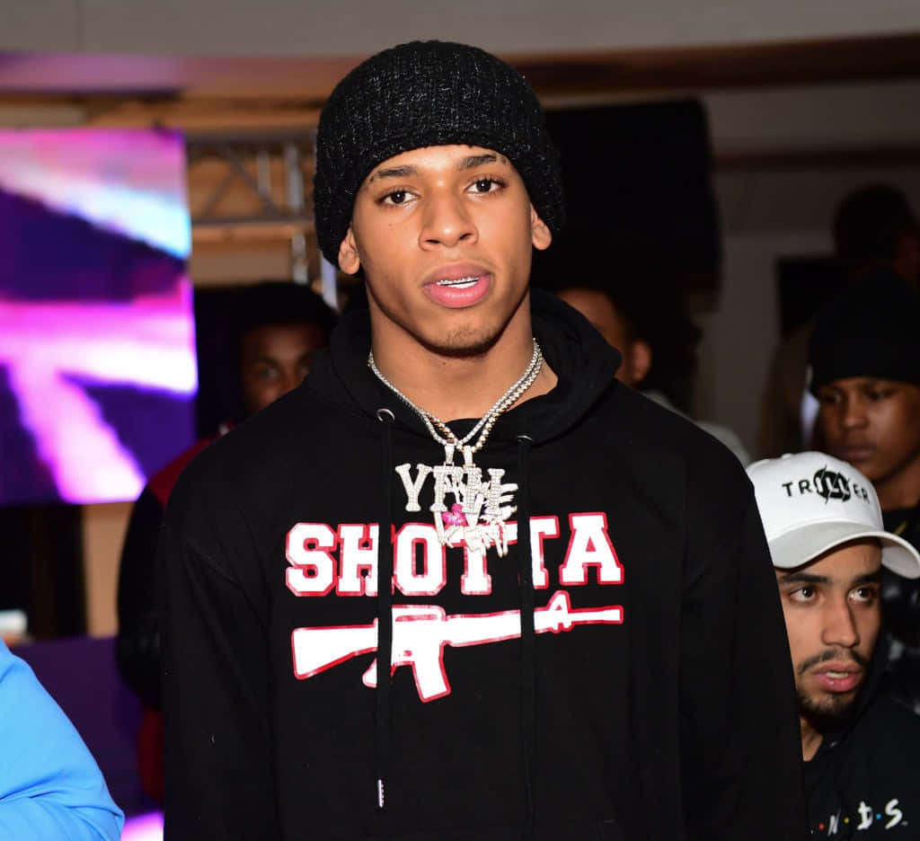 NLE Choppa 桌布 壁紙 — 一名戴著黑色毛帽、身穿印有「YALL SHOTTA」圖案連帽上衣的年輕男子，在擁擠的室內環境中直視鏡頭。
