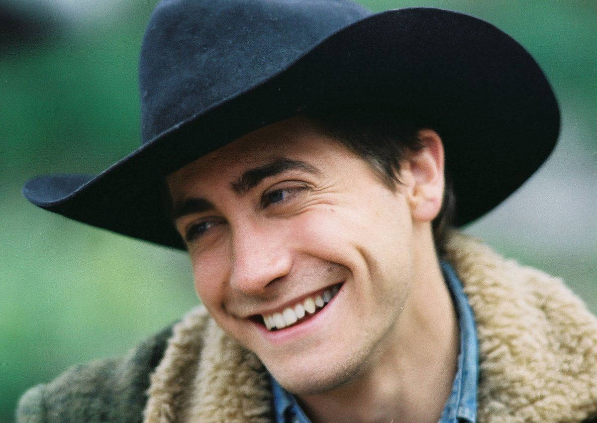 brokeback mountain 桌布 壁紙 — 一名戴著黑色牛仔帽的男子，身穿羊羔毛內裡外套，在柔和的綠色背景前露出燦爛的笑容。