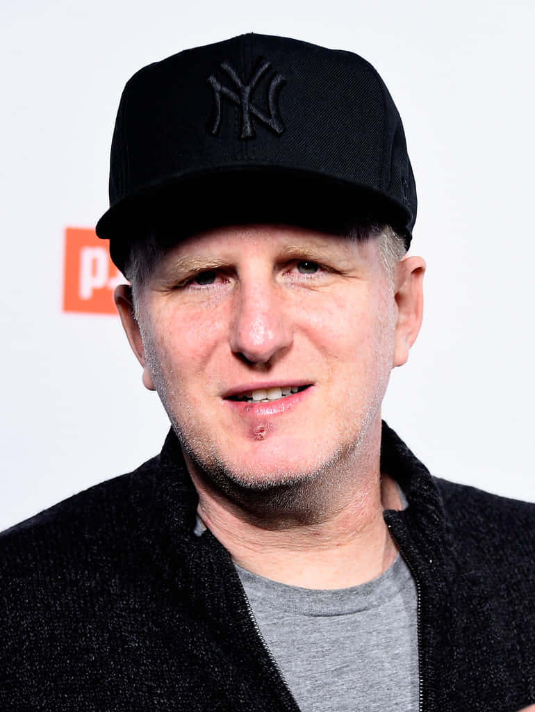Michael Rapaport 桌布 壁紙 — 一名戴著黑色紐約洋基隊帽子和深色外套的男子，在帶有紅色標誌的白色背景前微微一笑。