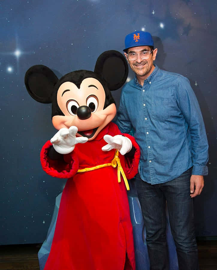 Ty Burrell 桌布 壁紙 — 一名戴藍色帽子的男子在星空背景下，微笑站在身穿紅色長袍的米奇老鼠吉祥物旁邊。