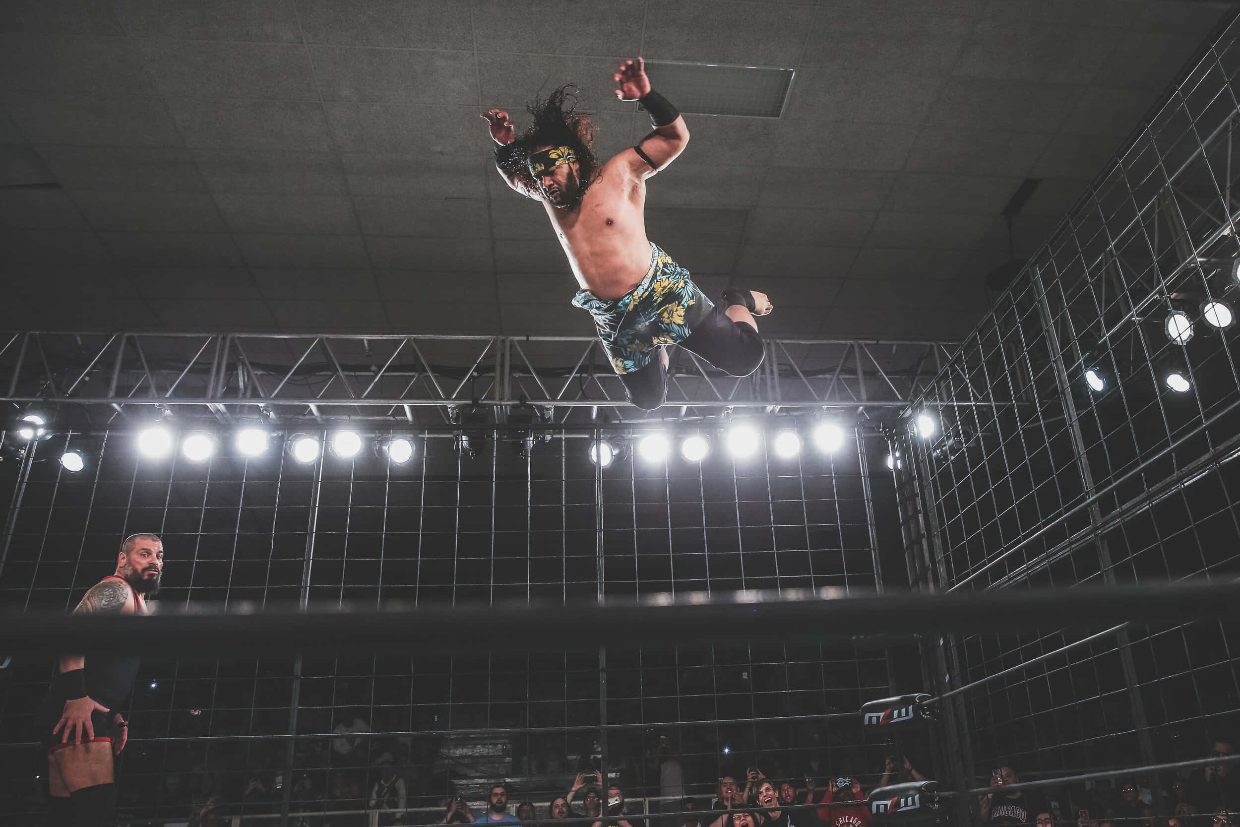 WWE 桌布 壁紙 — 一名戴面具的摔角手在明亮的場館燈光下，於摔角擂台上騰空躍起，下方有觀眾觀看。