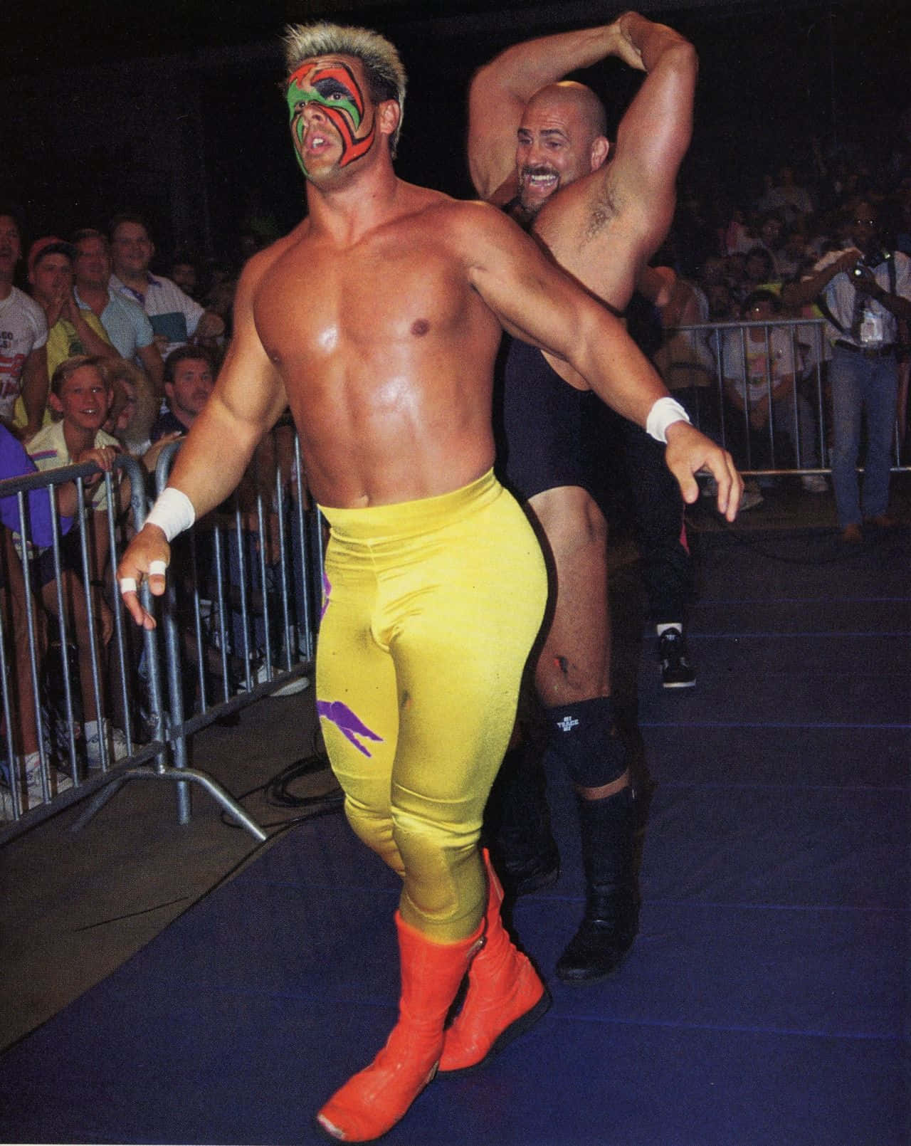 Nikita Koloff 桌布 壁紙 — 一名戴面具的摔角手身穿亮黃色緊身褲和紅色靴子，自信地大步走著，身後是一名光頭對手，場館座無虛席。