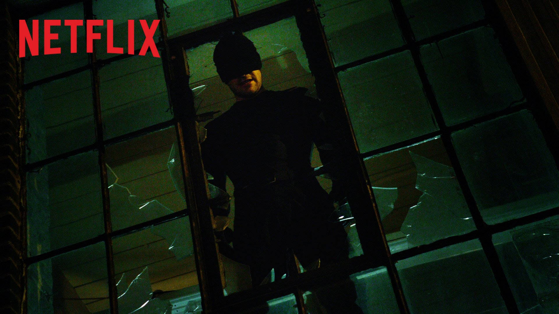 Netflix 桌布 壁紙 — 一名戴面具的義警站在破碎的窗戶後面，處於昏暗的城市環境中，帶有綠色調的燈光。