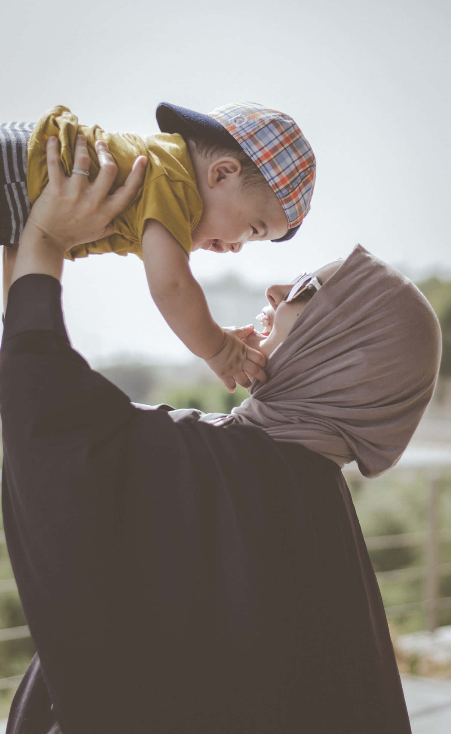 muslim 桌布 壁紙 — 一名戴頭巾的女性在明亮的戶外背景下，高興地舉起一個戴著格紋帽、微笑的幼兒。