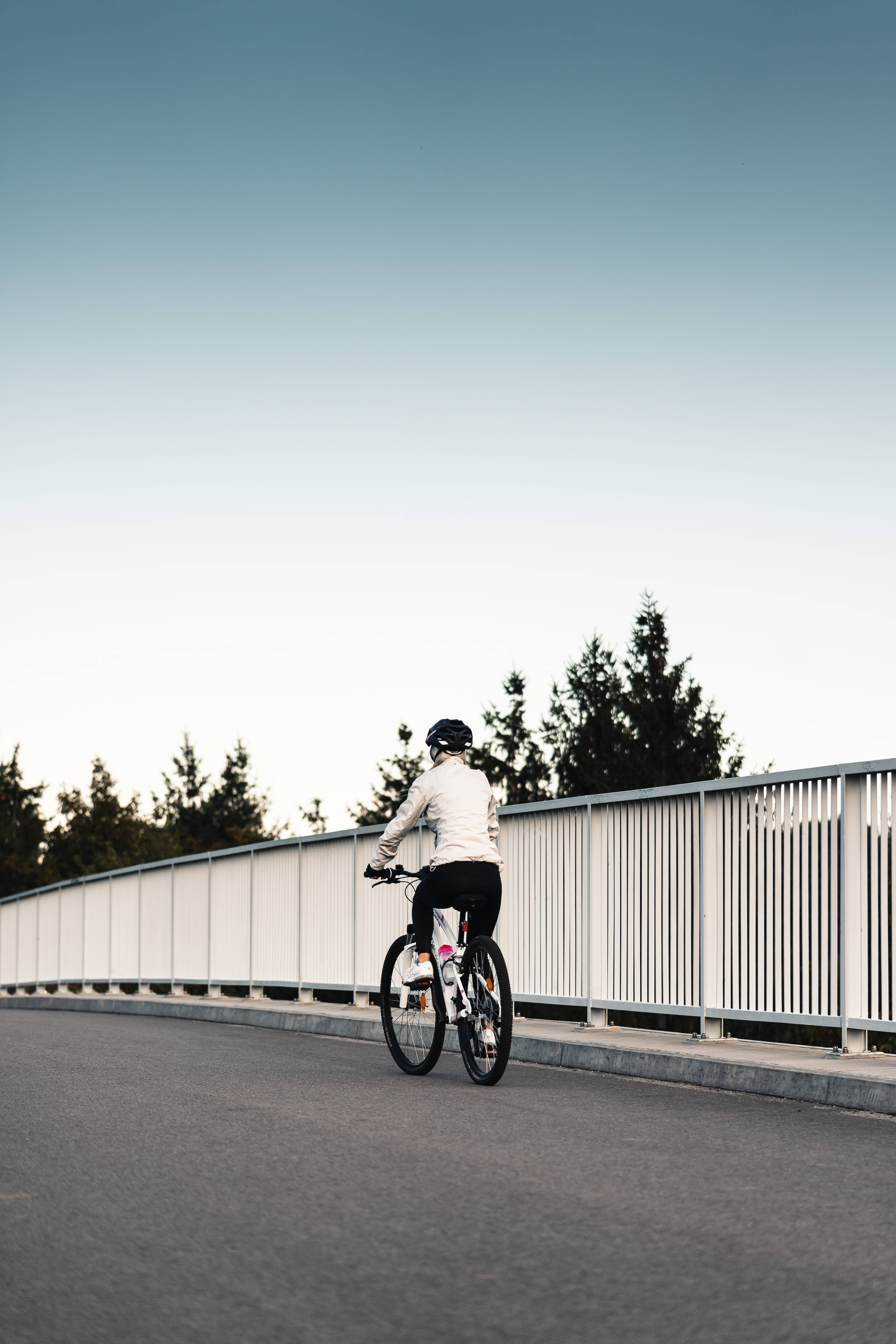 Bicycle 桌布 壁紙 — 一名戴頭盔、穿白色外套的自行車騎士在黃昏時分，在樹木和晴朗天空的背景下，踩著自行車駛離橋上。