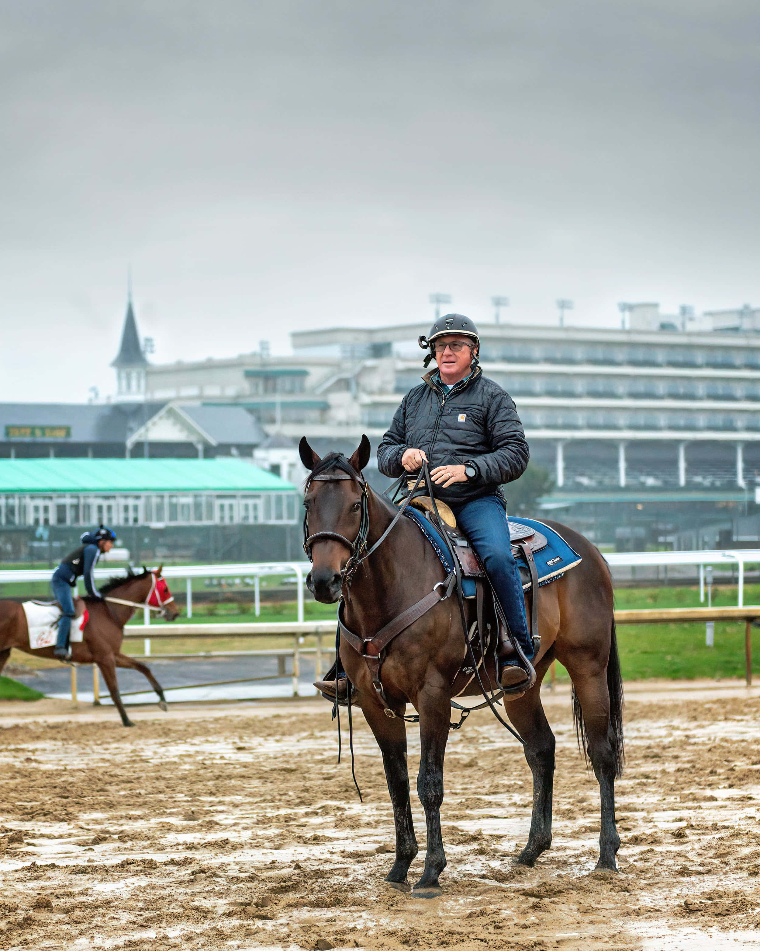 Kentucky Derby 桌布 壁紙 — 一名戴頭盔和穿外套的騎師坐在一匹棕色賽馬上，在泥濘的賽道上，背景有看台，天空多雲。