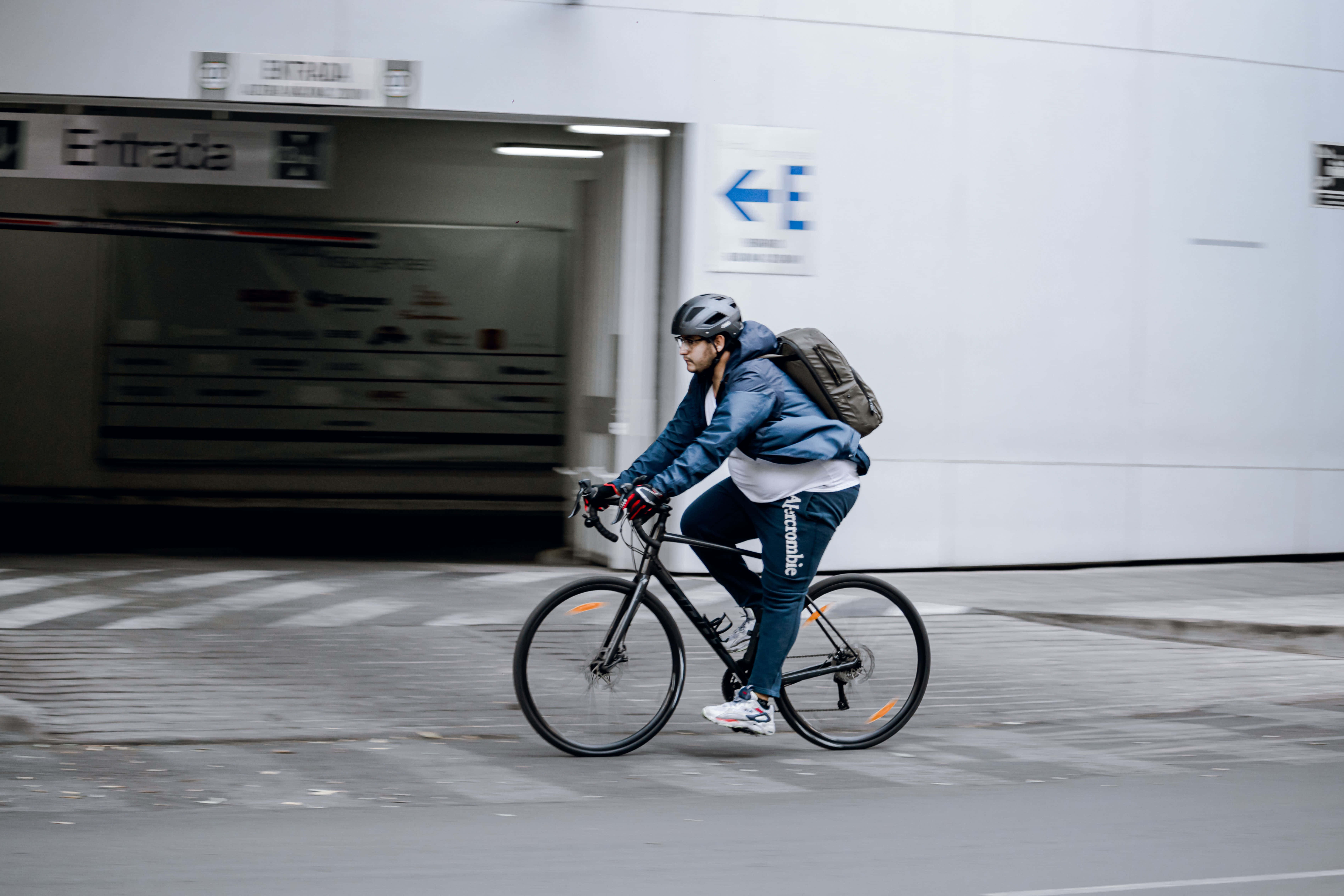 Navy Blue Jacket 桌布 壁紙 — 一名戴頭盔和背包的自行車騎士在都市人行道上騎過地鐵入口，帶有動態模糊效果。