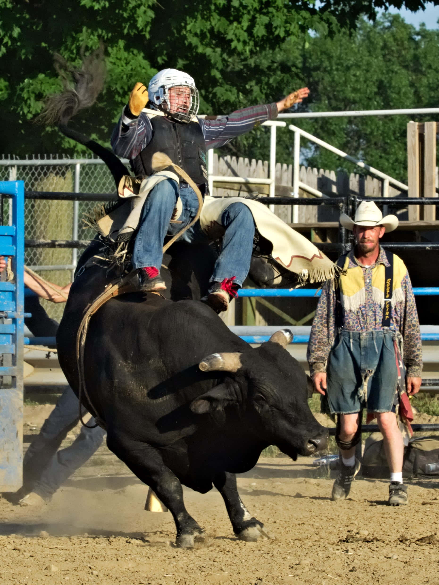 Bull Riding 桌布 壁紙 — 一名戴頭盔和背心的騎牛士緊抓著一頭騰躍的黑牛，牛仔競技場中塵土飛揚，附近有觀眾和馴牛師。