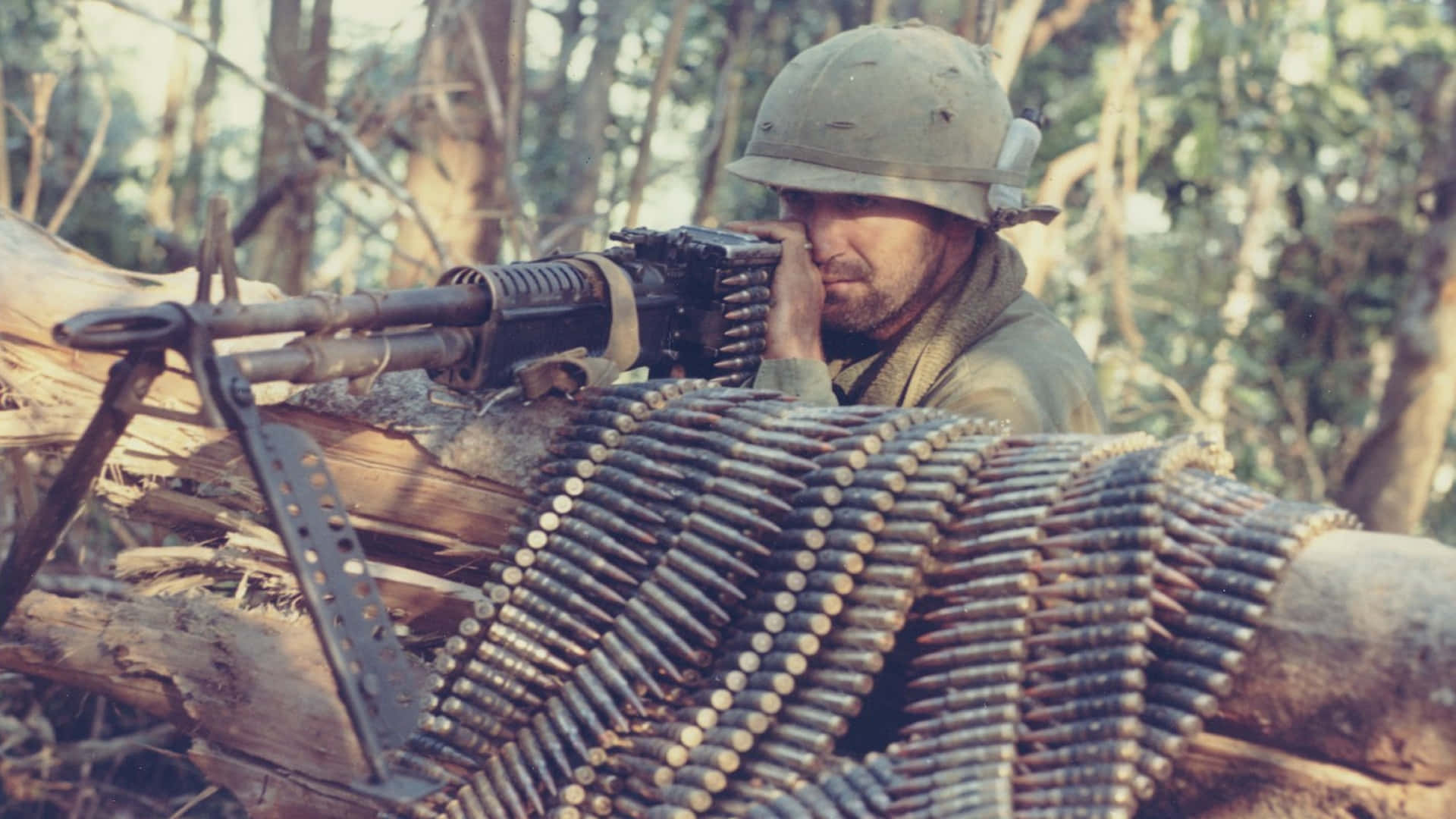 Vietnam War 桌布 壁紙 — 一名戴頭盔的士兵從樹林位置瞄準重型機槍，彈藥帶纏繞在圓木上。