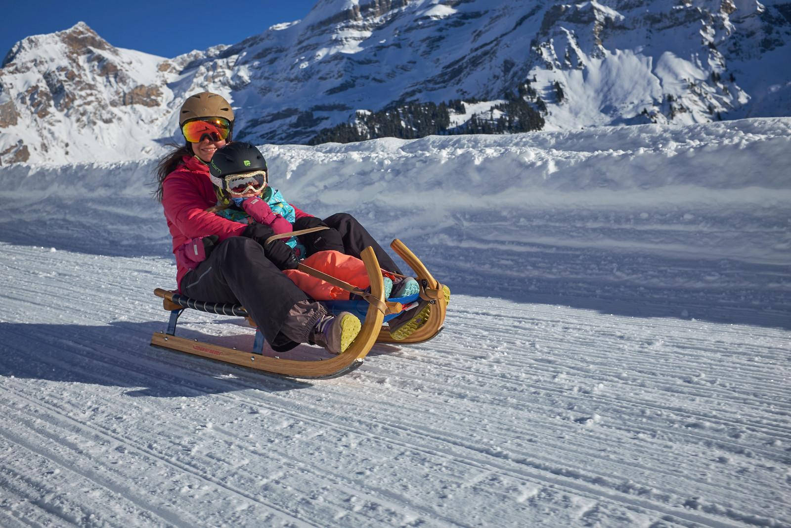  壁紙 — 一名戴頭盔的女子和小孩在陽光照耀的雪坡上滑雪橇，背景是白雪覆蓋的山脈。