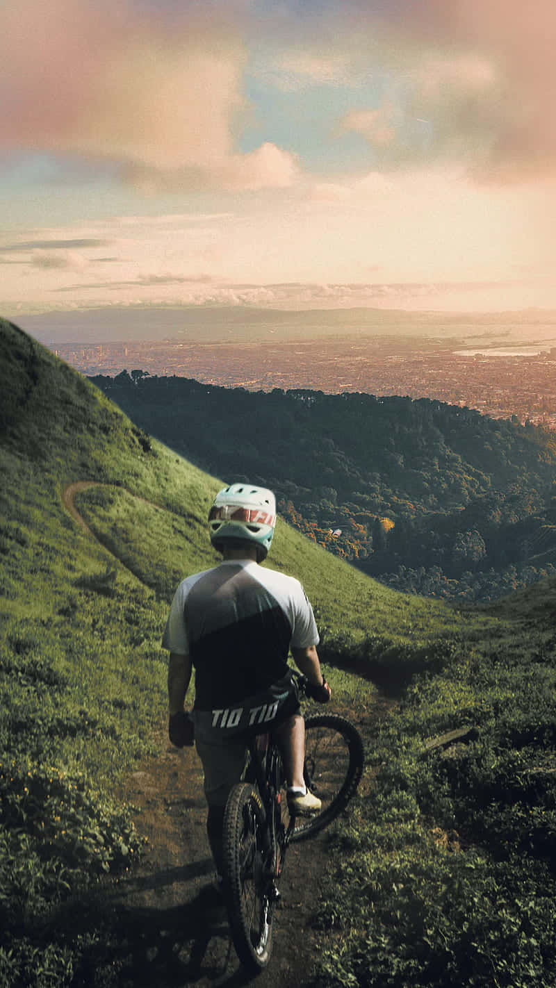 adventure 桌布 壁紙 — 一名戴頭盔的自行車手在日落時分停在長滿草的山頂上，俯瞰廣闊的城市山谷。