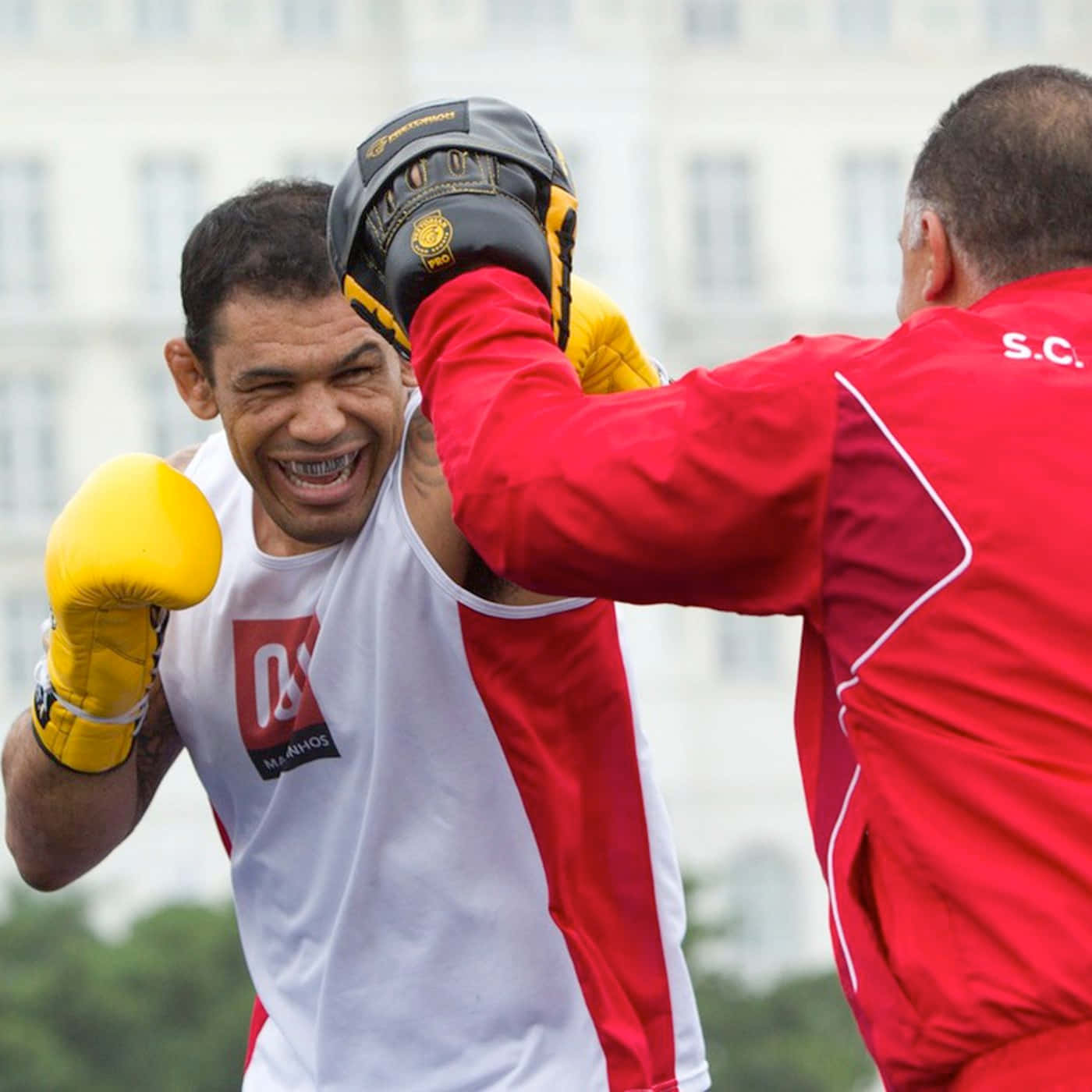 MMA 桌布 壁紙 — 一名戴黃色手套的拳擊手咧嘴而笑，他的紅衣教練在戶外訓練時出拳打他。