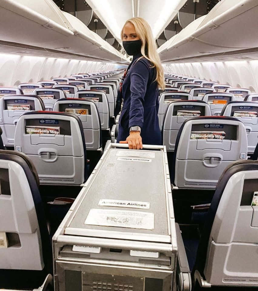 Aircraft 桌布 壁紙 — 一名戴黑色口罩的空服員推著手推車，沿著美國航空公司的飛機走道前進，兩旁是一排排空座位。